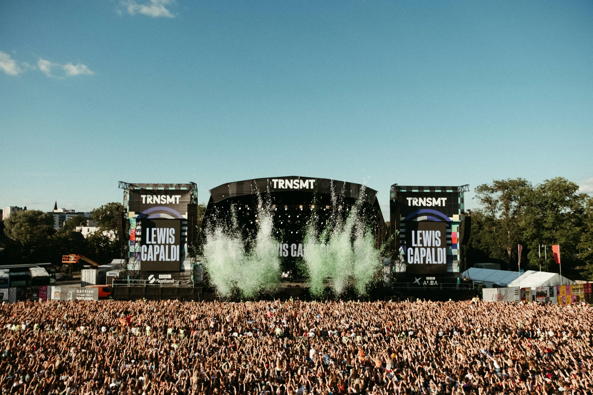 Large outdoor concert with a massive crowd, stage with screens displaying music artist Lewis Capaldi's name, and banners with "TRNSMT." Clear sky and cityscape in the background.