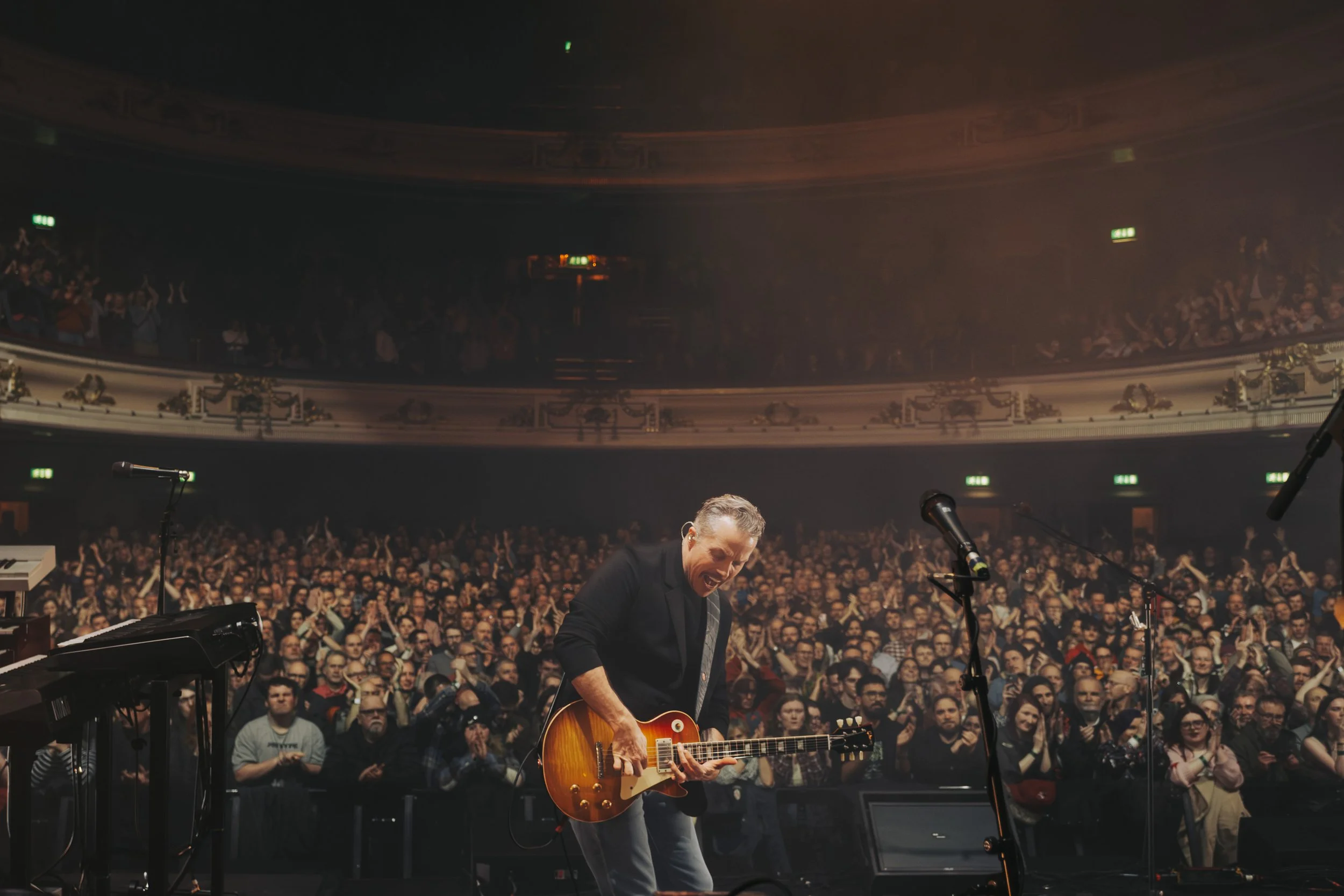 Musician playing guitar on stage with a crowd in the background, in a large theater with ornate interior and balcony.