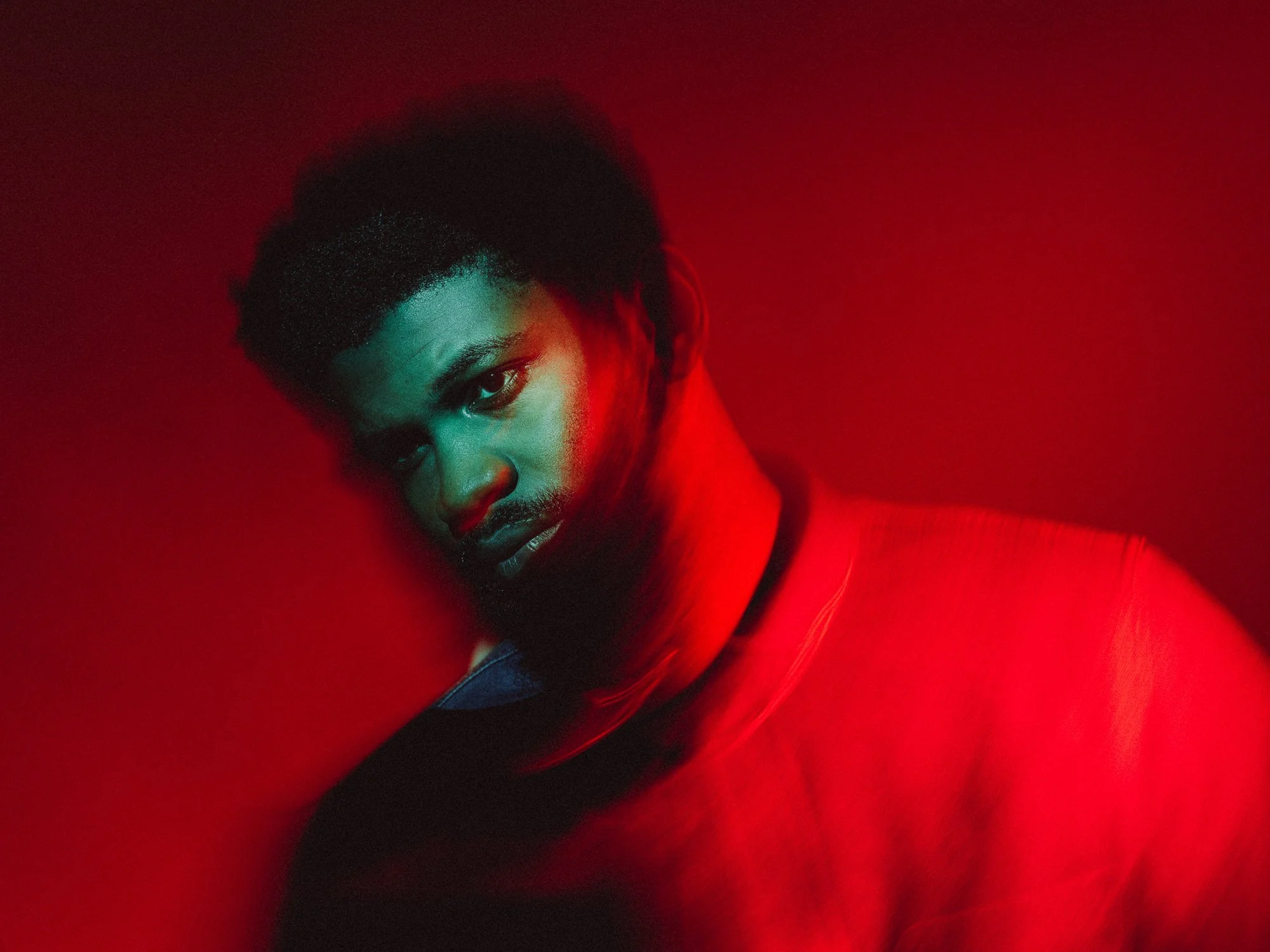 A close-up portrait of a person with curly black hair, intense gaze, and serious expression, lit with red lighting.