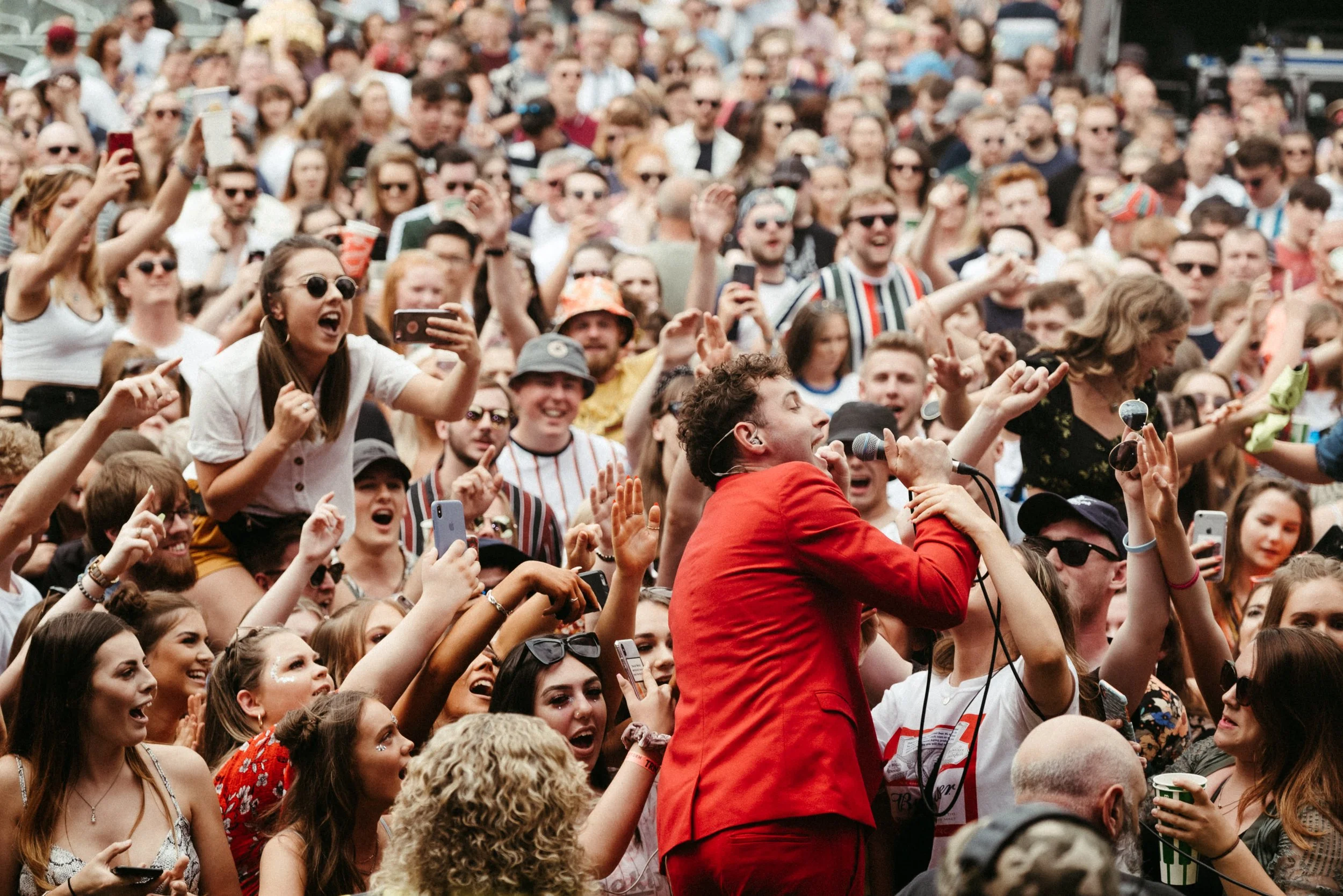 A large crowd of people at an outdoor event, many holding up phones and some wearing sunglasses, with a singer in a red jacket holding a microphone in the foreground.