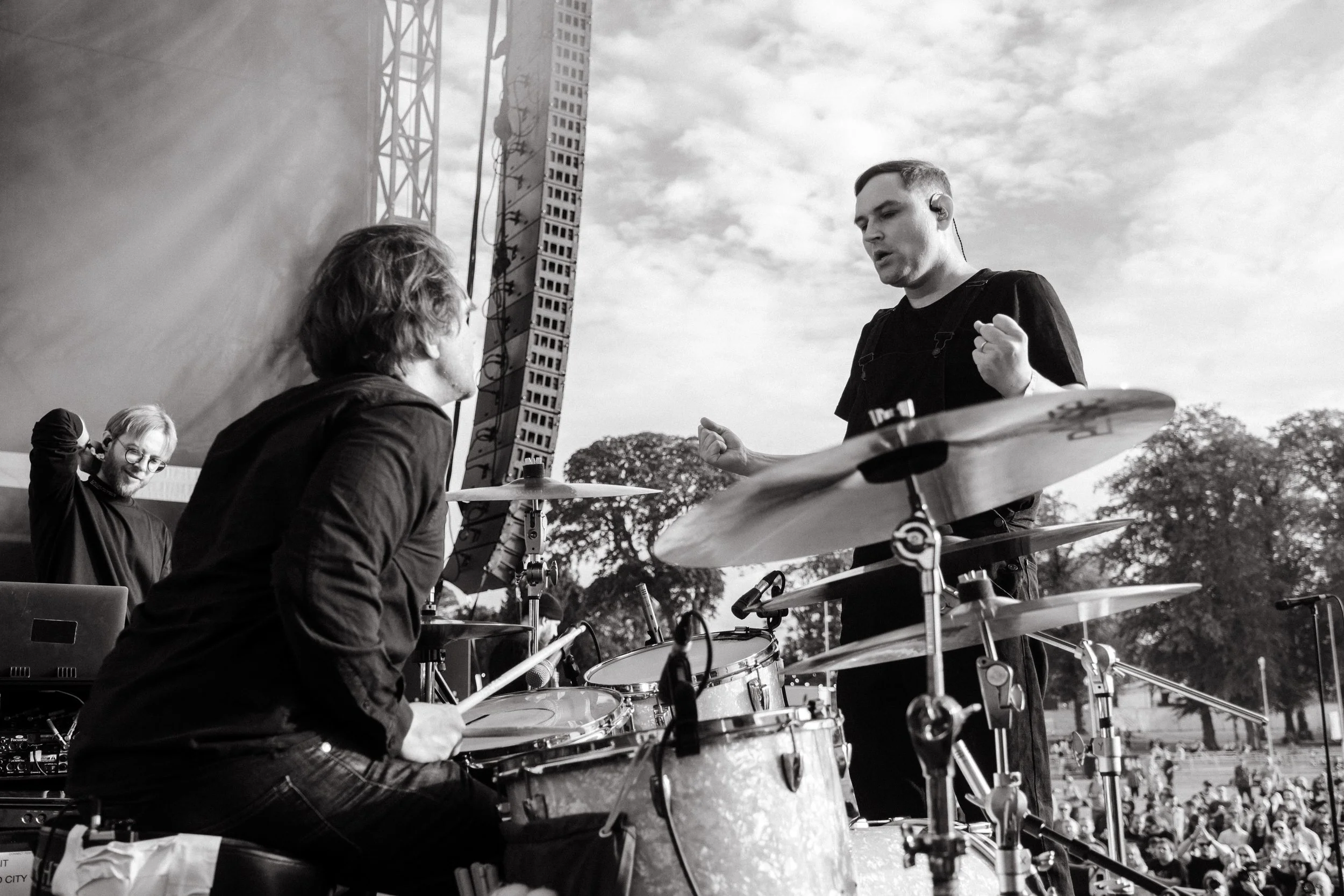 Black and white photo of musicians performing outdoors, with a man playing drums and another standing beside him, possibly conducting or speaking, with a crowd and trees in the background.