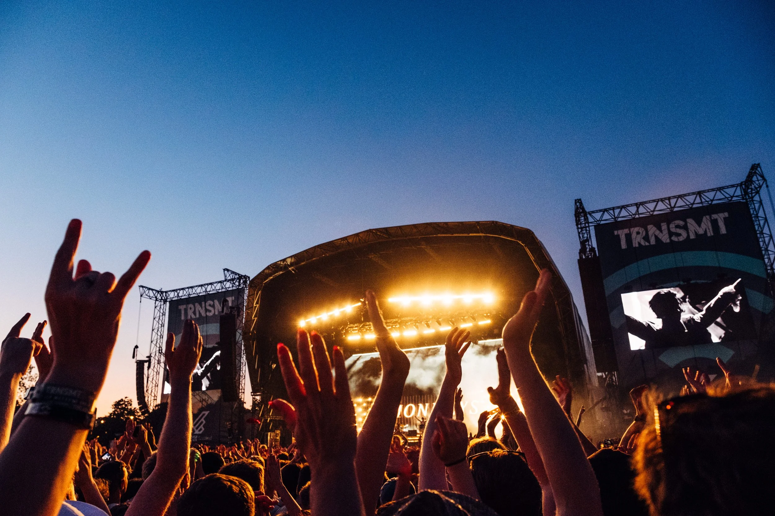 Crowd at an outdoor concert or festival during sunset, with hands raised and stage with bright lights and screens in the background.