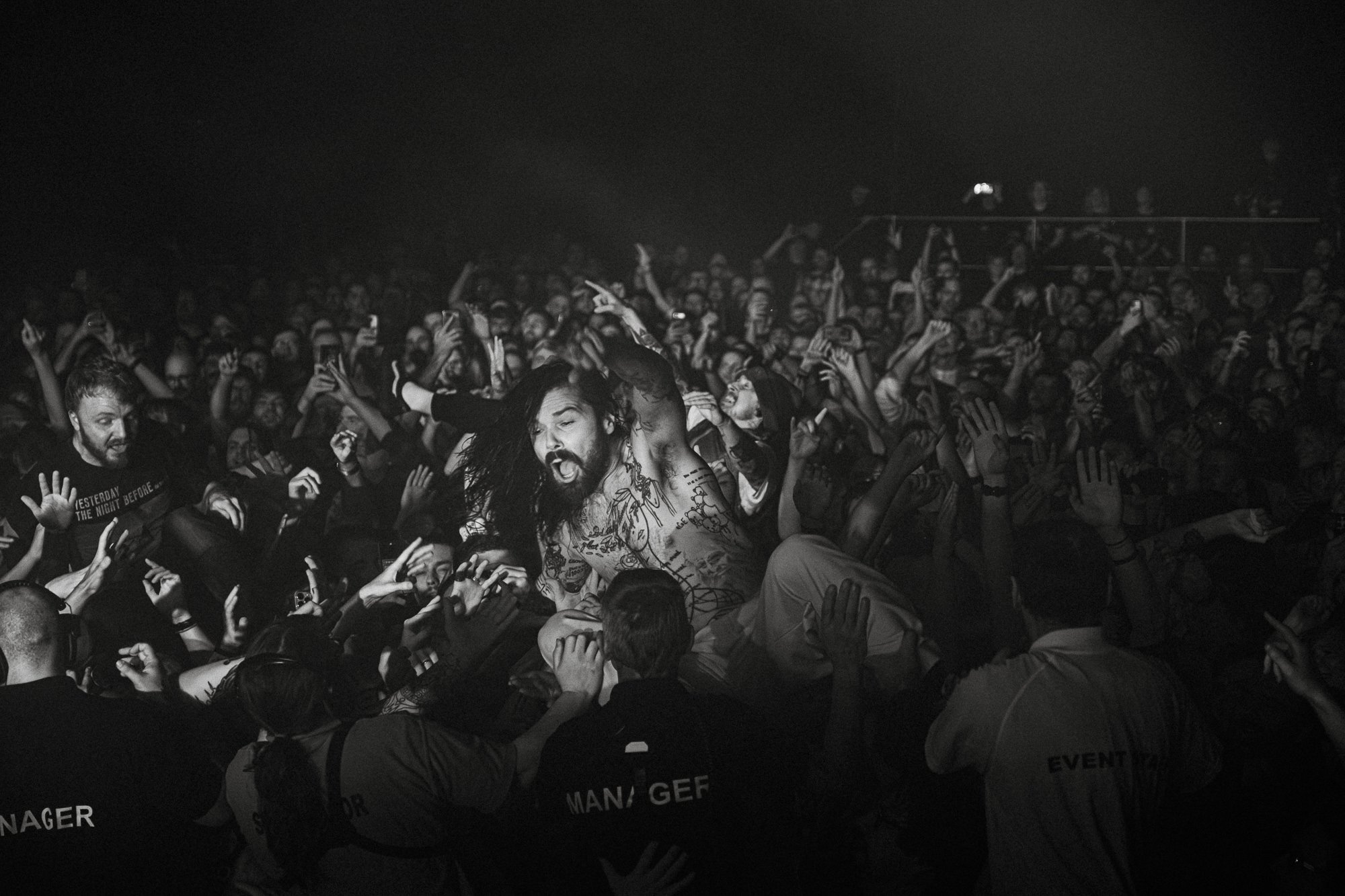 A black-and-white photo of a large crowd at a concert or event. The crowd is actively engaged, with many people reaching out and taking photos or videos. In the center, a shirtless individual with long hair, tattoos, and facial hair appears to be per