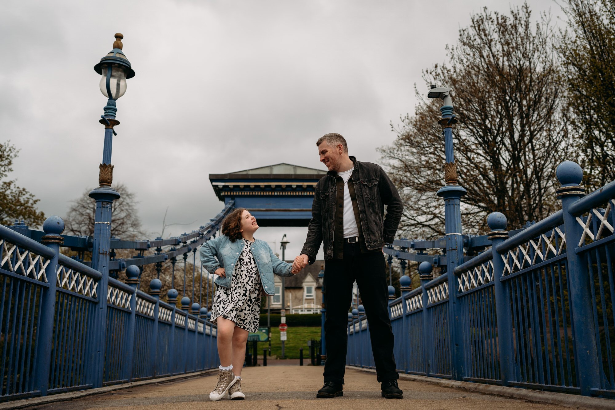 A man and a young girl are holding hands and smiling on a bridge in an outdoor park, with cloudy skies and trees in the background.