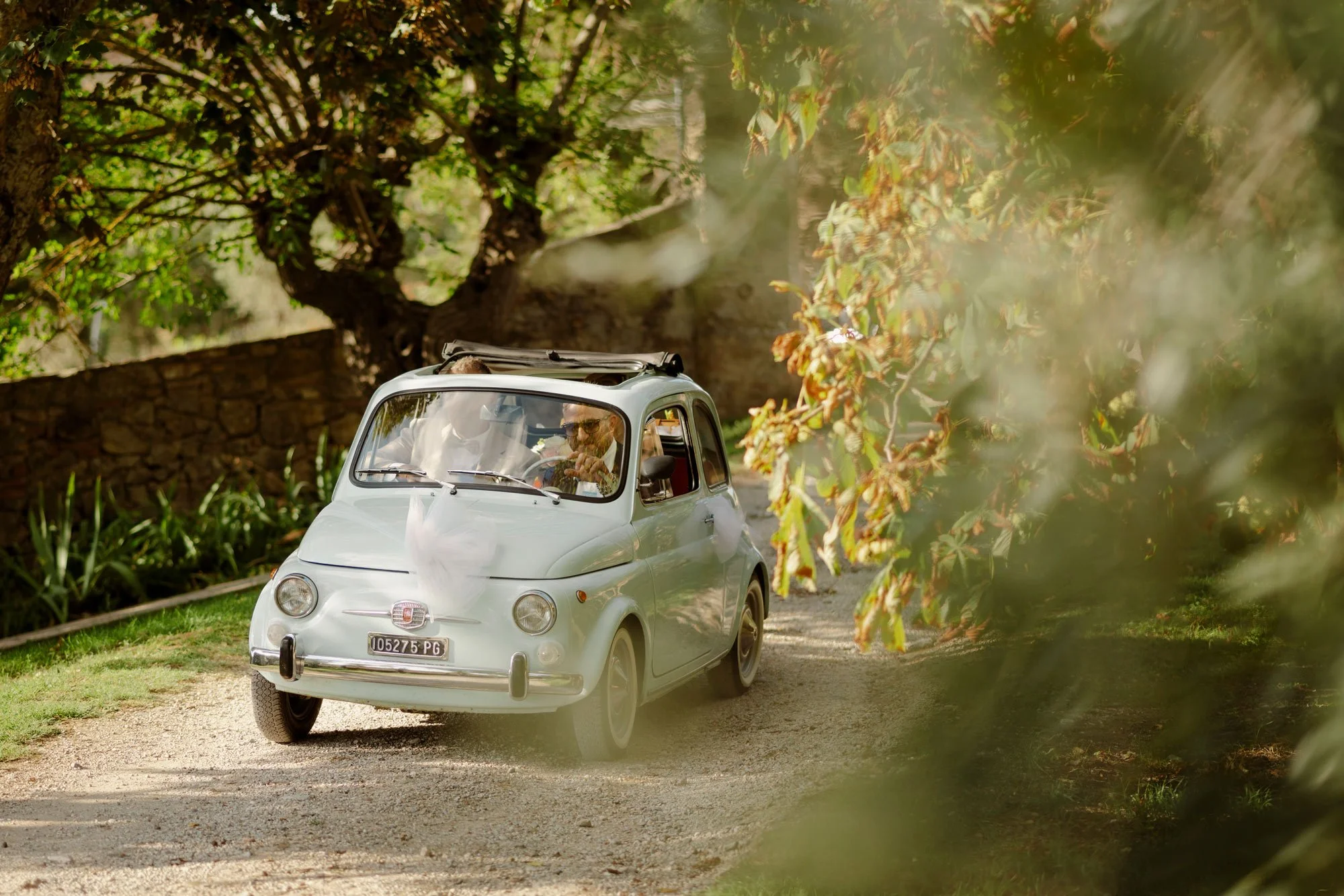 En liten hvit retro Fiat 500 kjører på en grusvei omgitt av grønne trær og busker.
