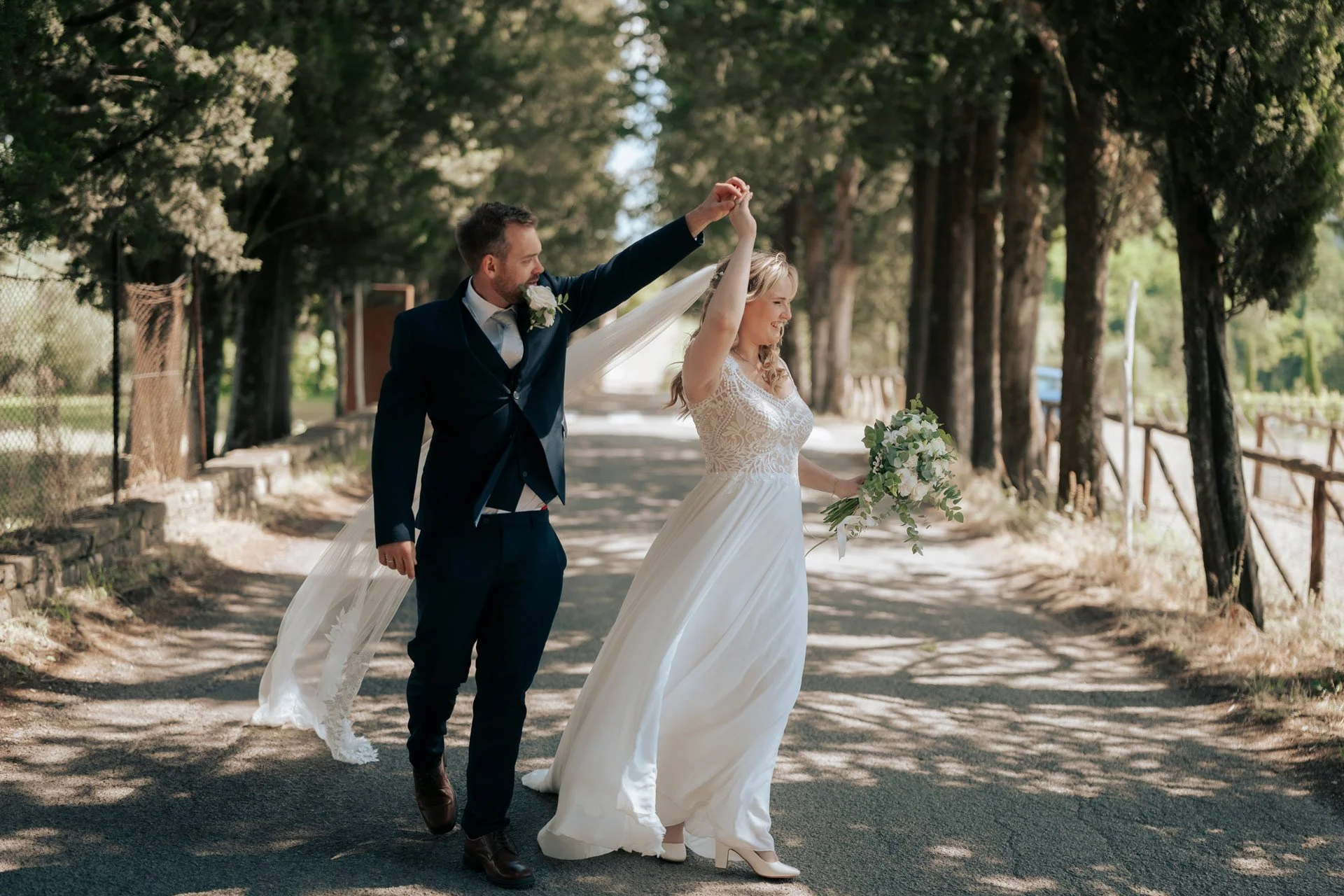 Et brudepar bestående av en mann og en kvinne danser på en grusvei omgitt av trær. De er kledd i flotte klær, med kvinnen i en lang, hvit brudekjole og mannen i en dress.
