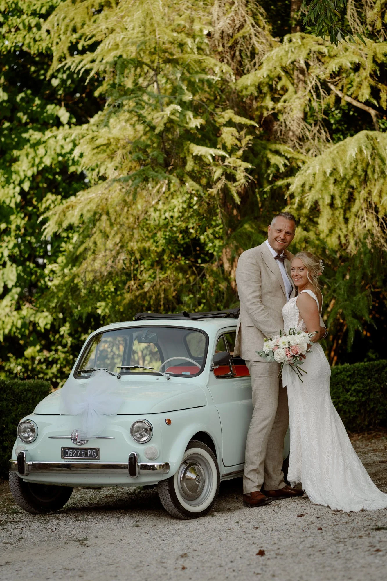 Et nygift par står foran en vintage hvit bil med blomsterdekor, omgitt av grønne trær og busker i en utendørs setting i Toscana