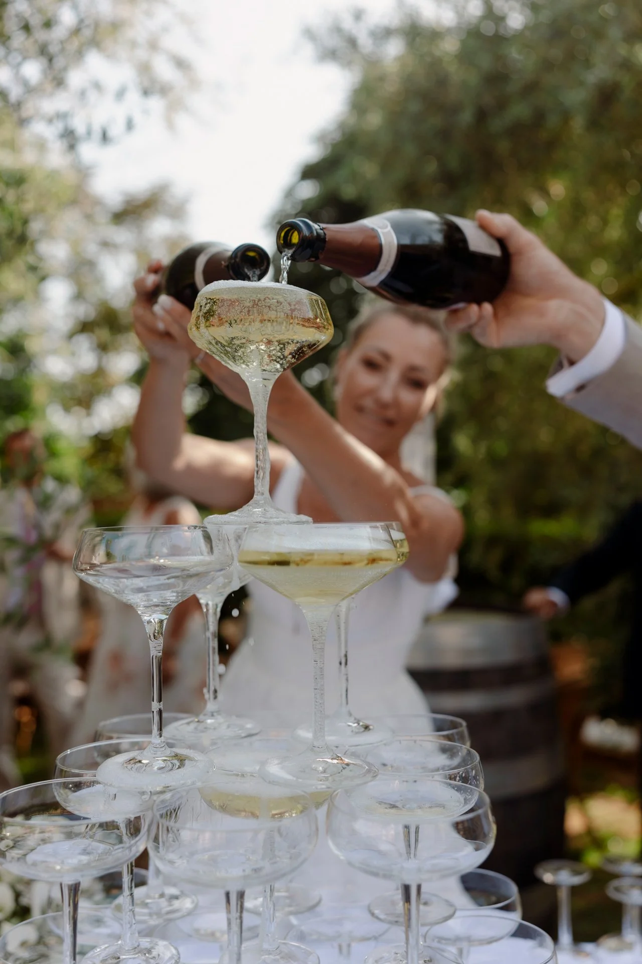 En kvinne i brudekjole fyller et glass med champagne fra en flaske, første rad med flere tomme champagneglass synlige, utendørs med trær i bakgrunnen.