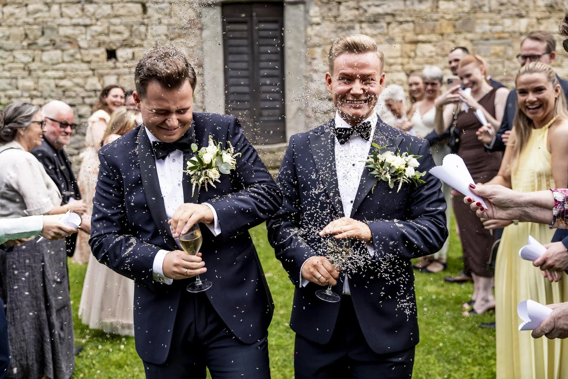 To nygifte menn i selskapsdress kommer gående med champagne i hånden, omringer av glade mennesker under en bryllupsfeiring utendørs.