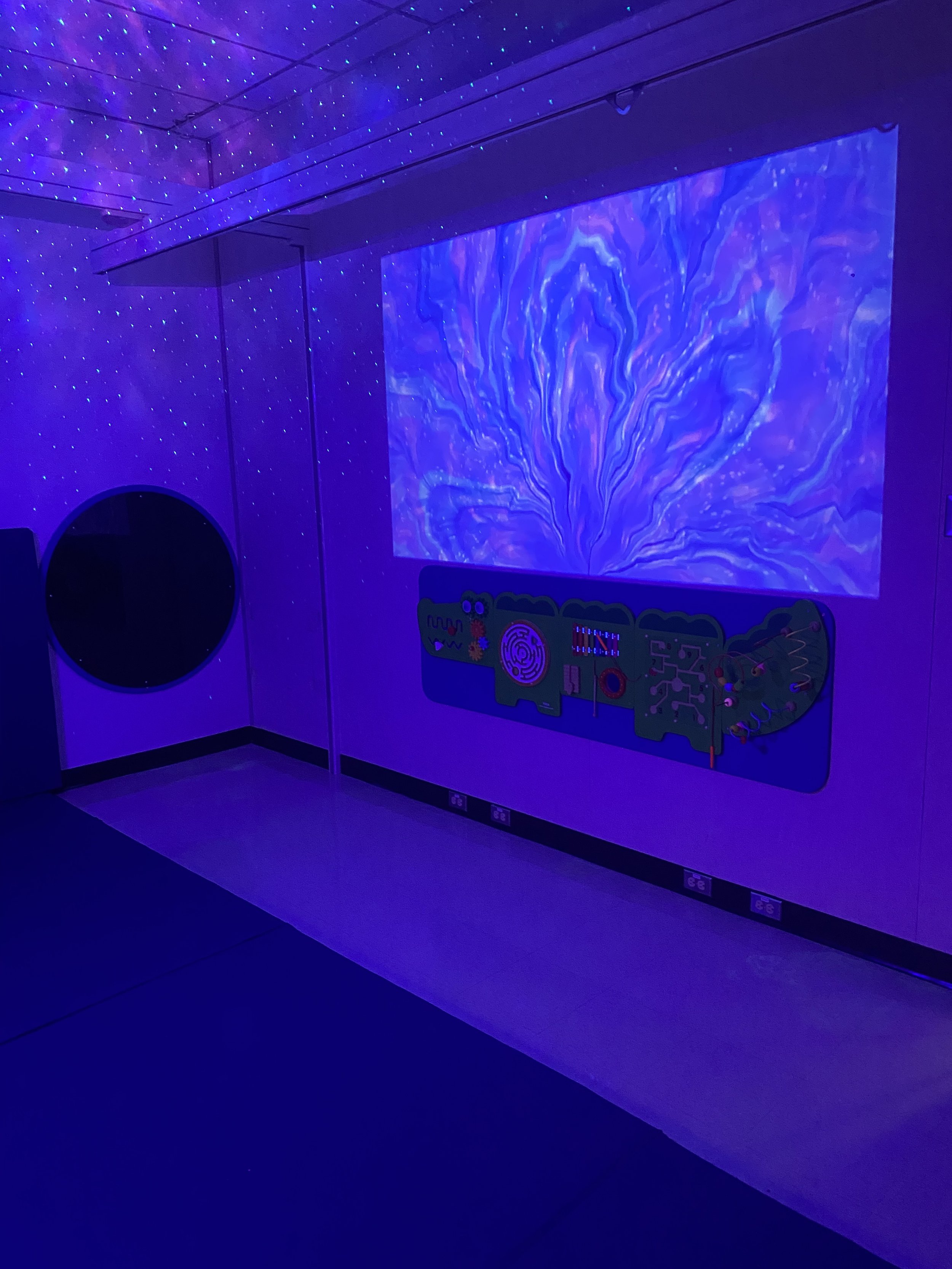 A room decorated with black lights, with a large digital projection of a blue and purple abstract pattern on the wall. There are small blue lights resembling stars on the ceiling, and a circular opening or mirror on the wall. A control panel with various buttons and knobs is mounted on the wall below the projection.