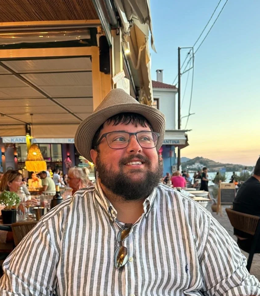 A man with glasses, a beard, and a striped shirt wearing a straw hat, sitting outdoors at a restaurant during sunset with water and hills in the background, surrounded by other diners.