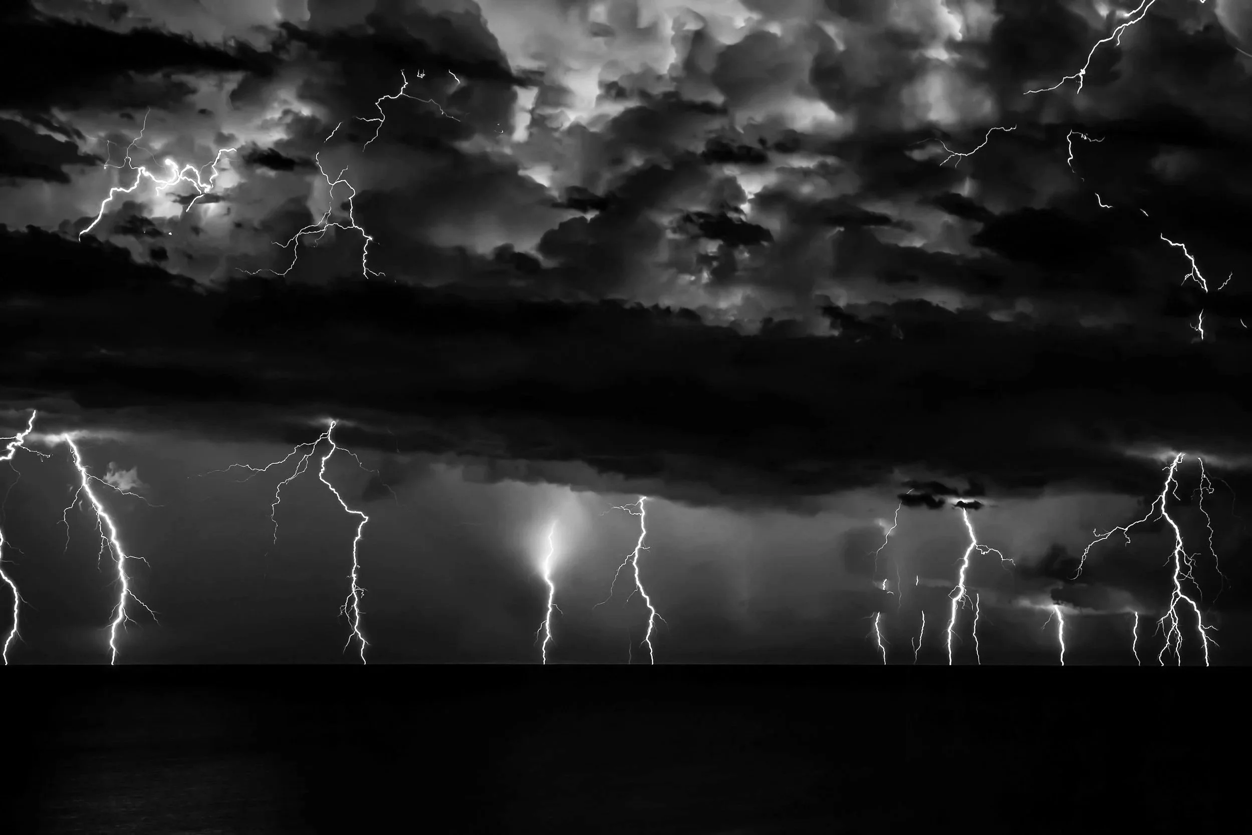 Black and white photo of a stormy sky with numerous lightning bolts striking down over a body of water.