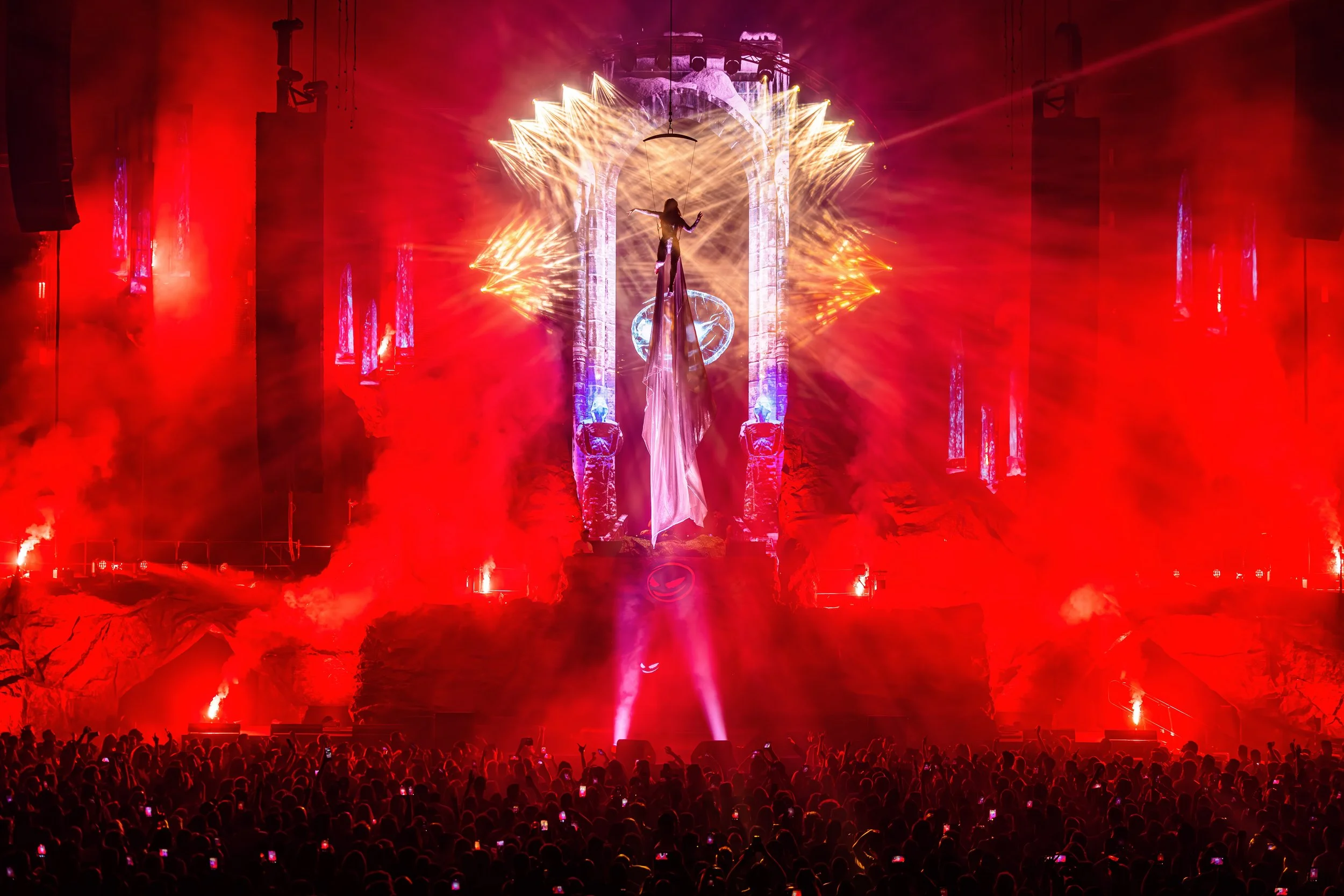 Een live concert met rood verlichte lichten, een aerialist in een lange jurk hangt in tissue boven een groot publiek dat omhoog wijst of foto's neemt.