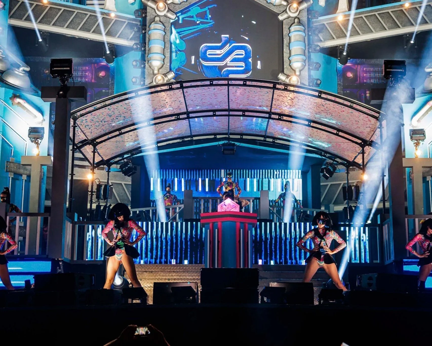 Een grootschalig festival met mainstage dansperformance op een futuristische stage met heldere blauwe en paarse verlichting, dansers op het podium in kleurrijke kostuums.