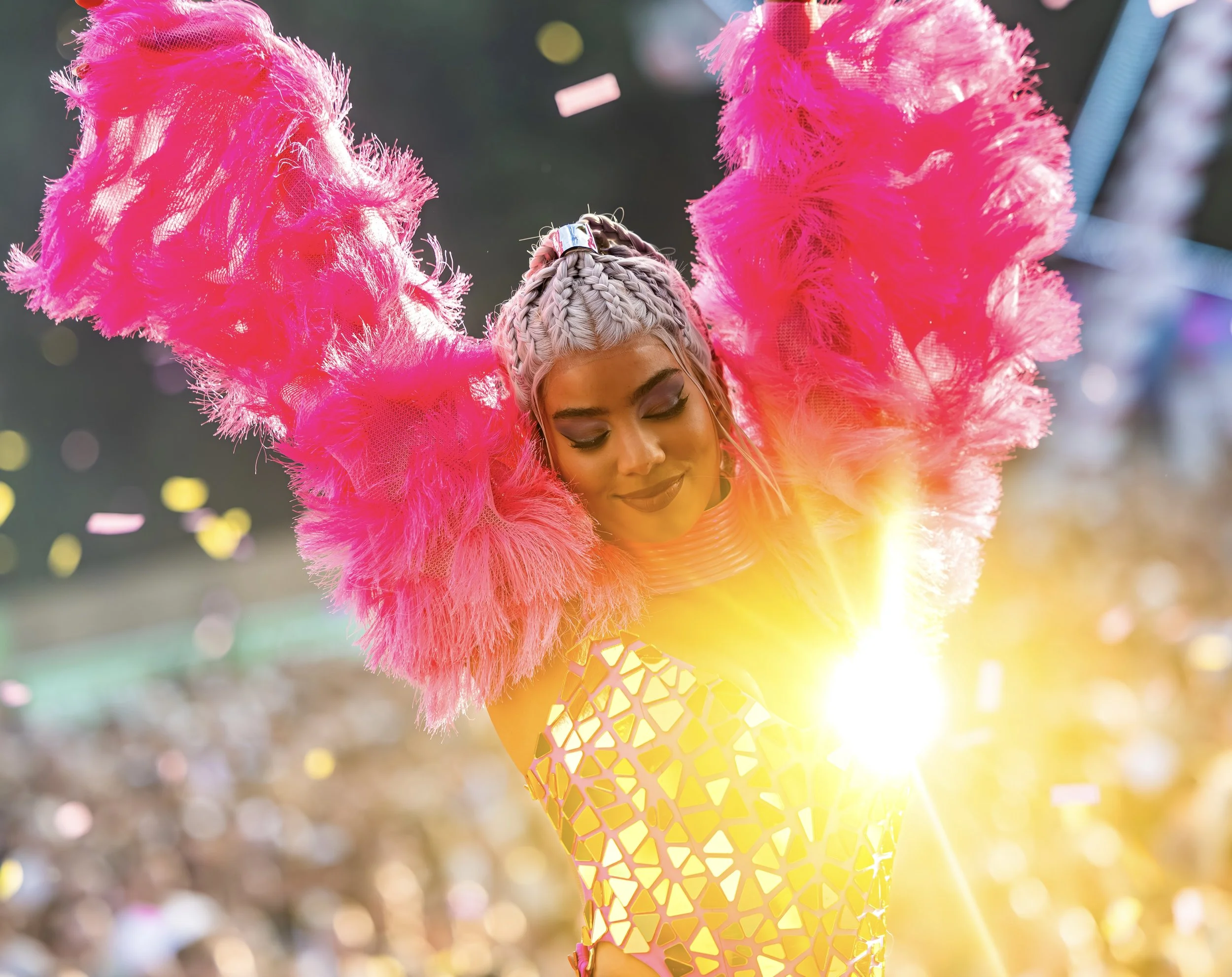 Vrouw in kleurrijk feestoutfit met grote roze veren aan haar schouders, lachend met gesloten ogen, zonsondergang en confetti op de achtergrond.