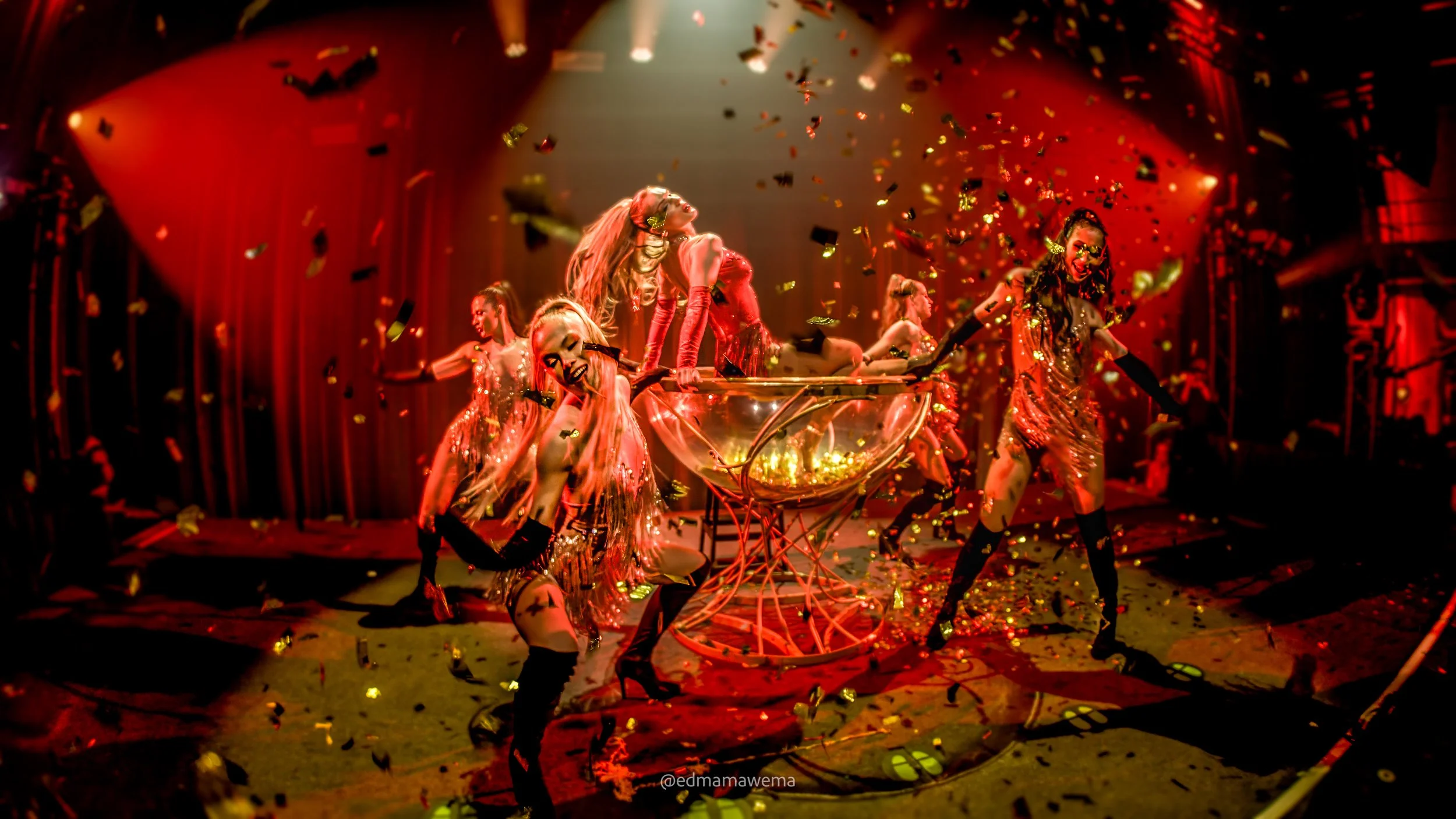 Dansers vieren een feestje met confetti en rode verlichting, gekleed in glitterende outfits op een podium.