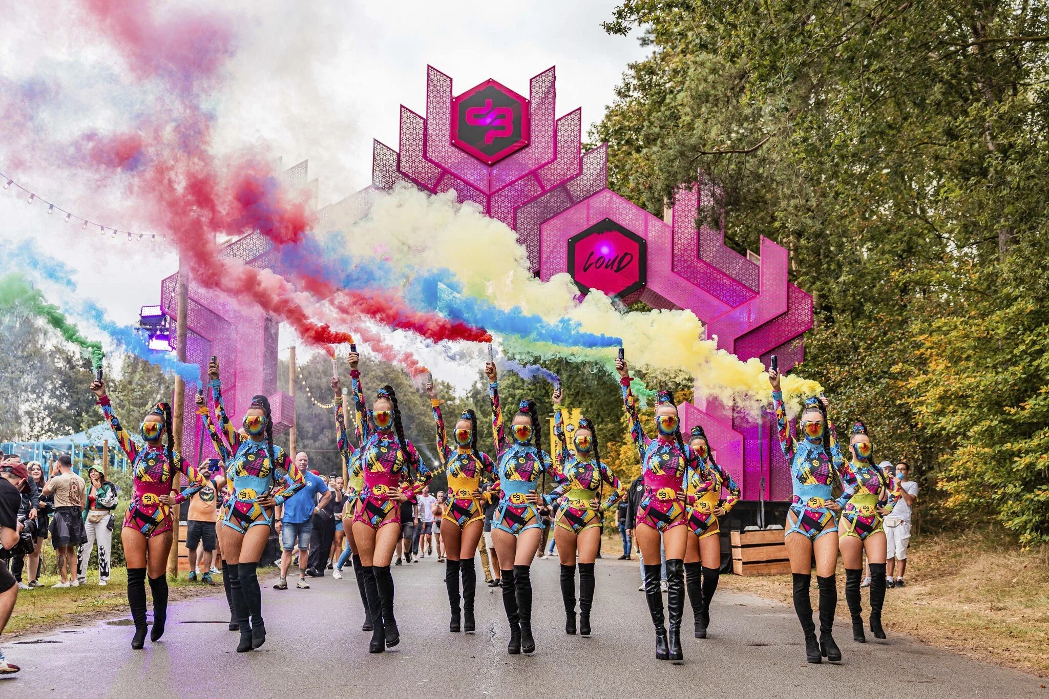 Vrouwen in kleurrijke outfits en gele maskers marcheren met hand omhoog tijdens festival met pink decor en kleurrijke rook, omringd door publiek en groene bomen.