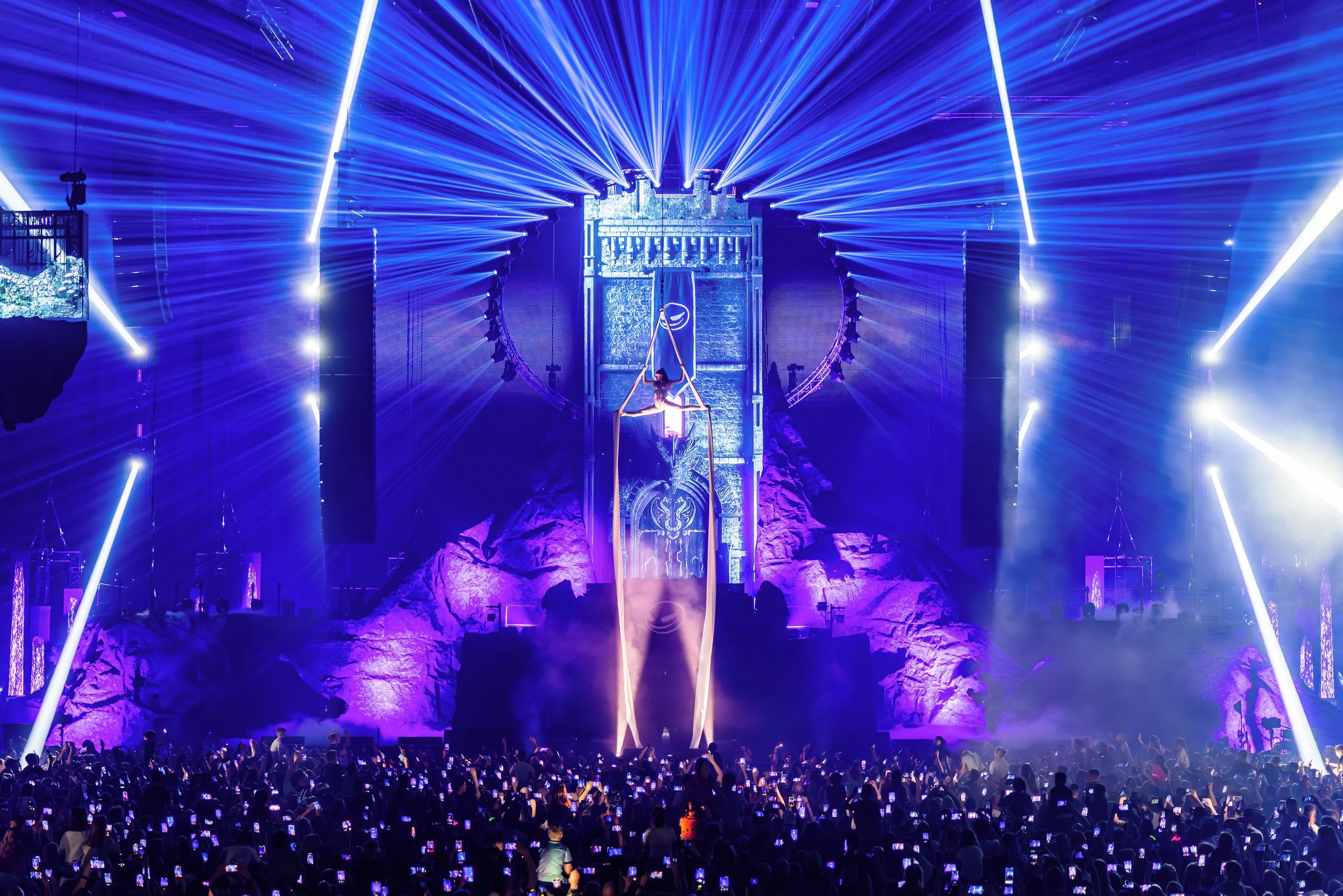 Grote live entertainment show met aerialist in tissue boven een grote massa mensen met lichtshow, lasers en publiek tijdens een evenement of festival.