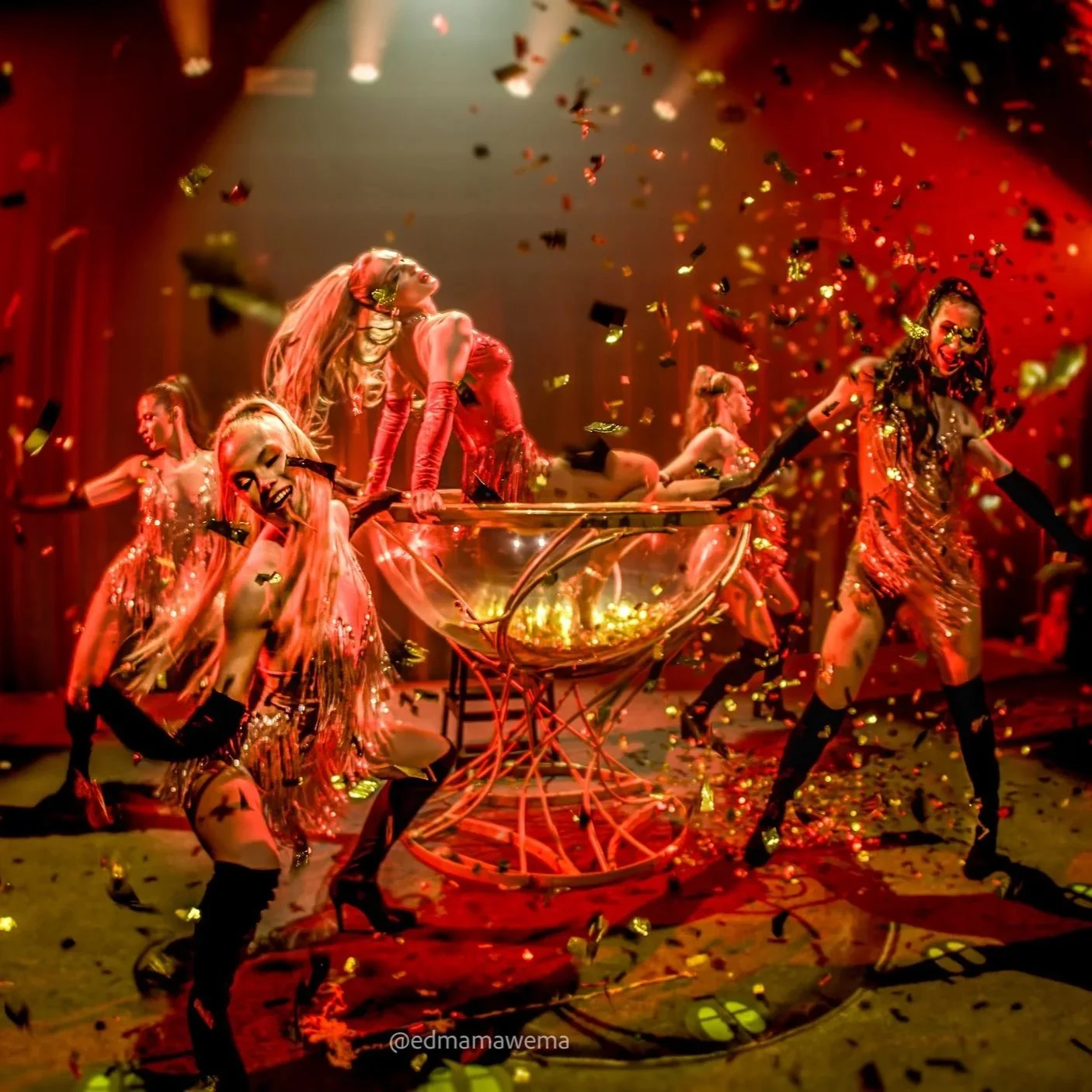 Vijf showgirls op een podium dansen rondom een groot champagne glas omringd door gouden confetti, dansen en lachen, met rode en gouden verlichting op de achtergrond.