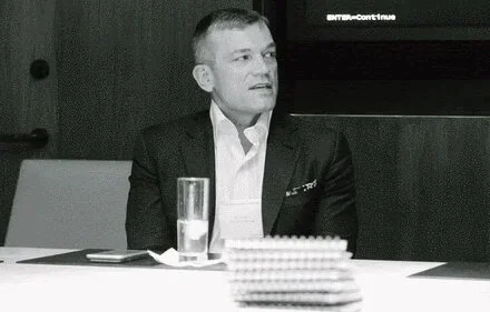 A man in a suit sitting at a table with a glass of water and a notebook.