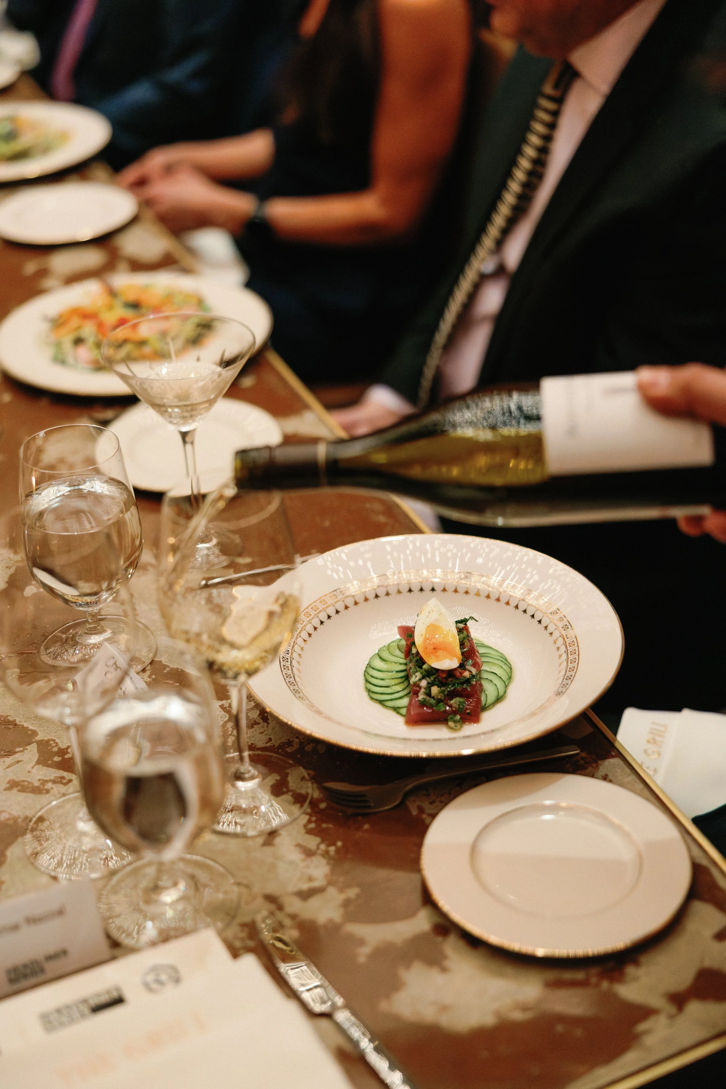 Elegant restaurant table setting with plates of food, glasses of water, and a person pouring wine, featuring a dish with cucumber, tomato, and a poached egg.