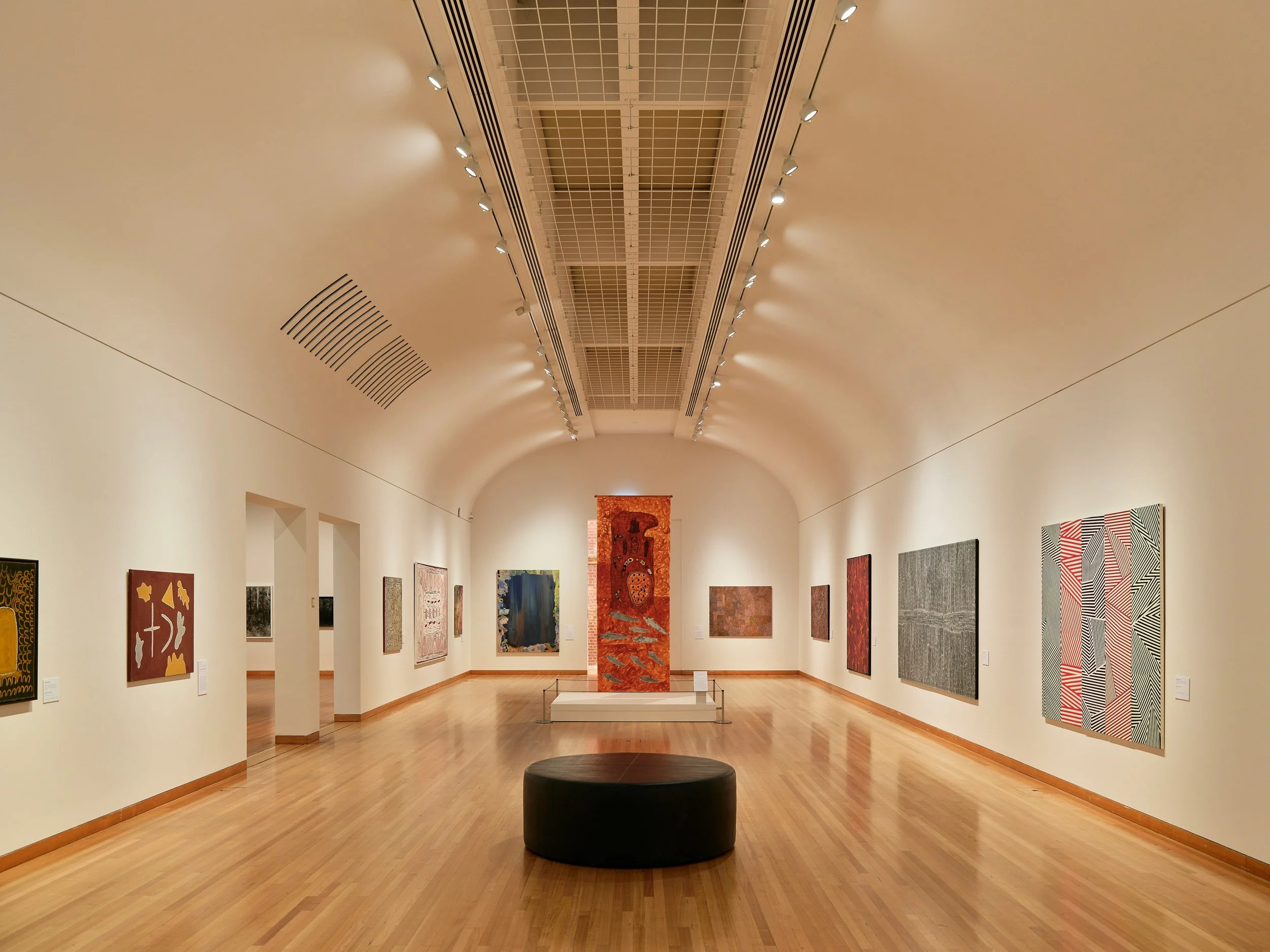 An art gallery with colorful abstract paintings on white walls, a wooden floor, and a black circular seating area in the center.