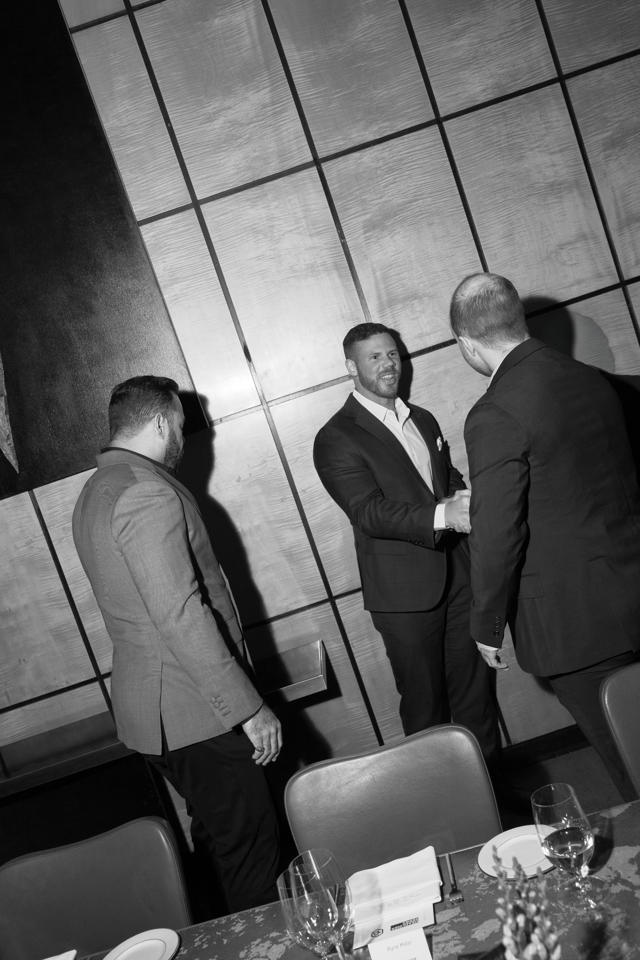 Three men in formal attire are conversing at a dinner event. Two are facing each other, shaking hands, while the third stands nearby. The background features a wooden wall with a geometric pattern, and the table in the foreground is set with glasses, plates, and napkins.