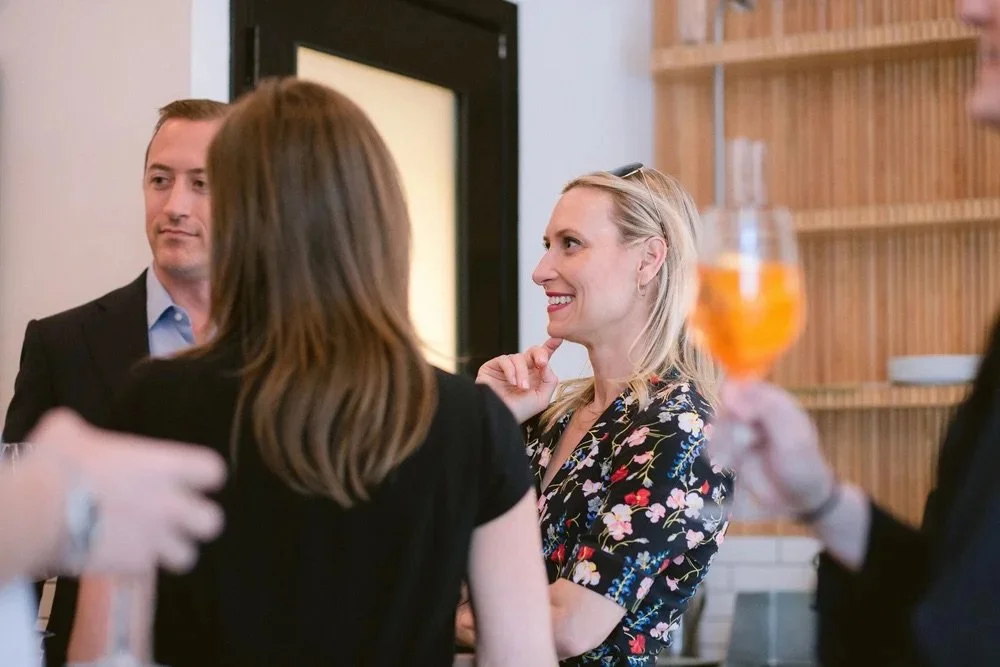 People socializing at a gathering, with a woman in a floral dress smiling and talking, and a person holding a glass of beer.