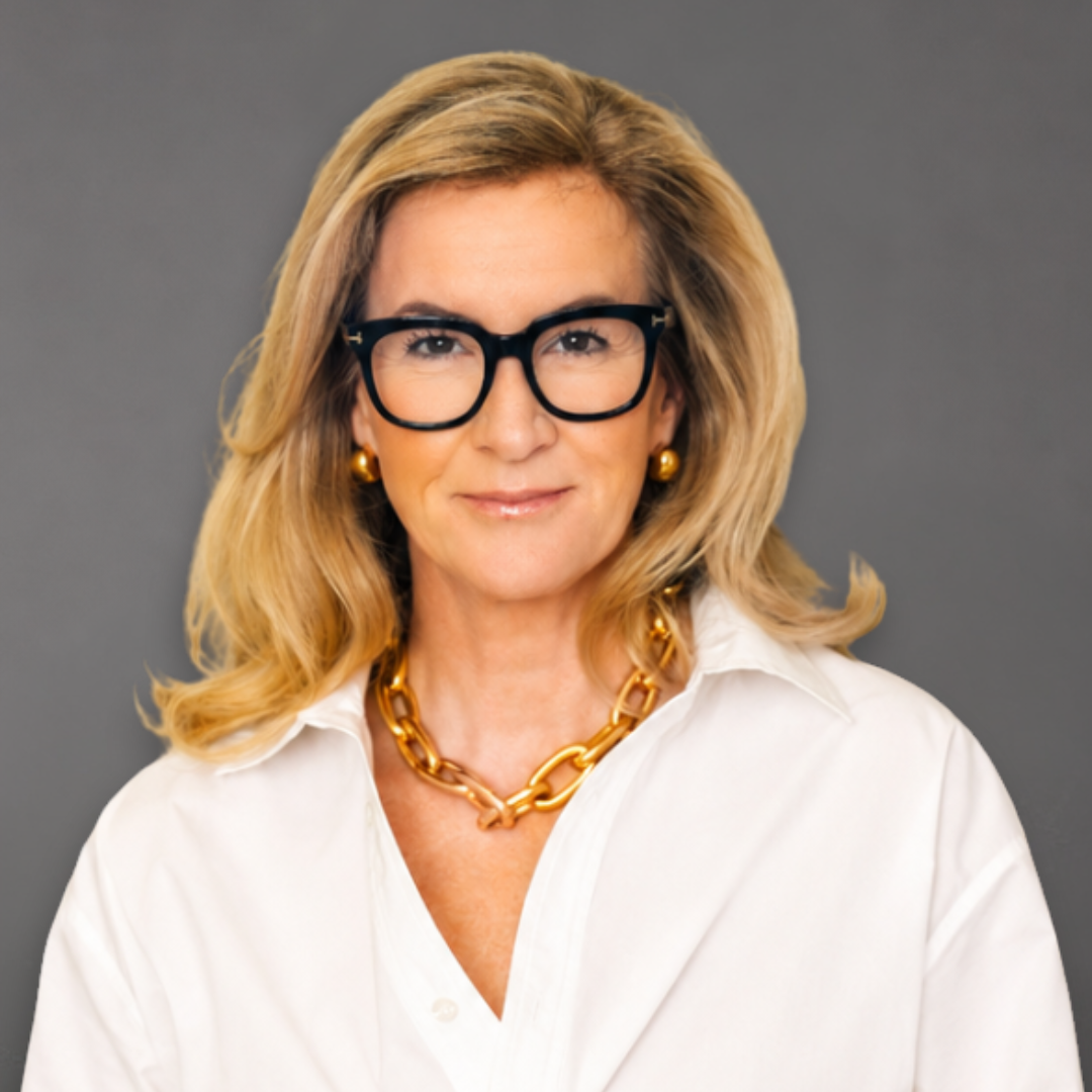 A woman with blonde hair, wearing black glasses, gold earrings, a gold chain necklace, and a white blouse, smiling and leaning against a white wall.
