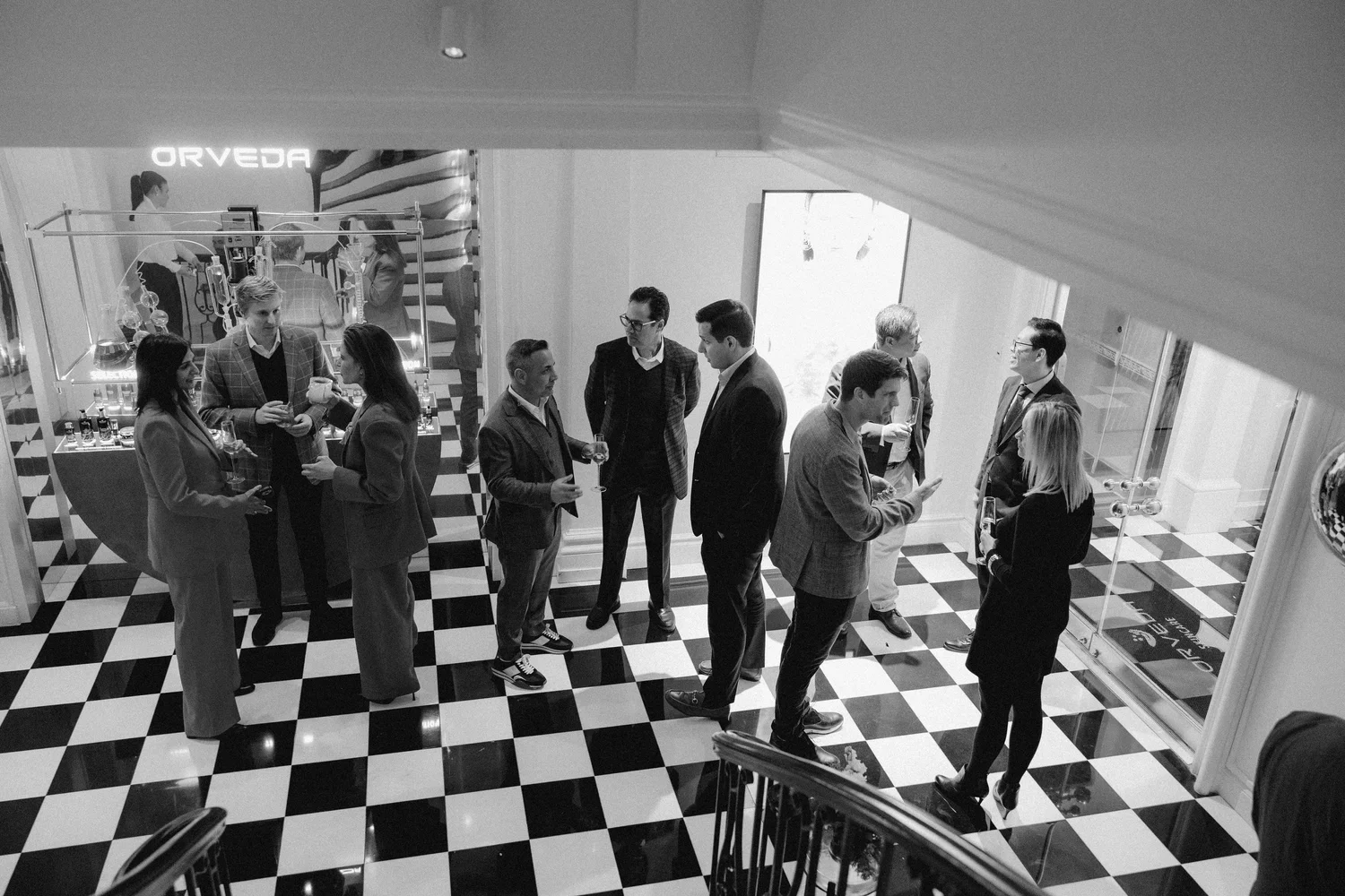 A group of people at a social gathering in a stylish indoor space with checkered floor, engaged in conversation and holding drinks.