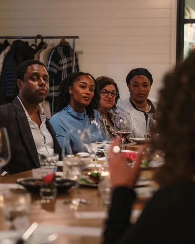 Group of people sitting at a dining table during a dinner event, with plates, glasses, and food in front of them.
