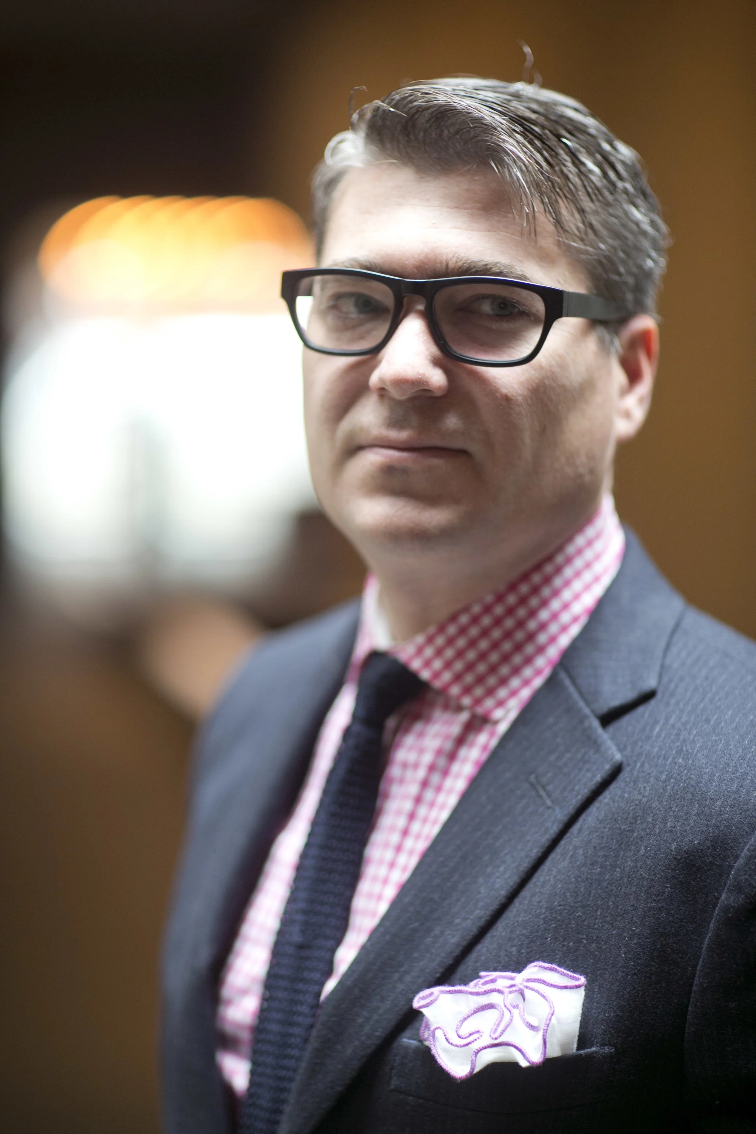 Close-up of a man with glasses, wearing a dark suit with a pink and white checkered shirt and a folded pocket square with pink and white ruffles in the breast pocket.