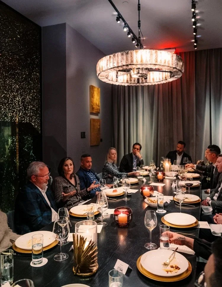 A group of people sitting around a long formal dining table in a dimly lit room with modern decor and candles.