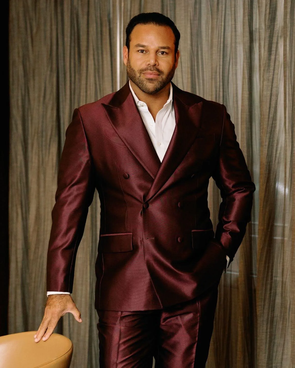 A man wearing a dark red suit with a white shirt, standing in front of a curtain.
