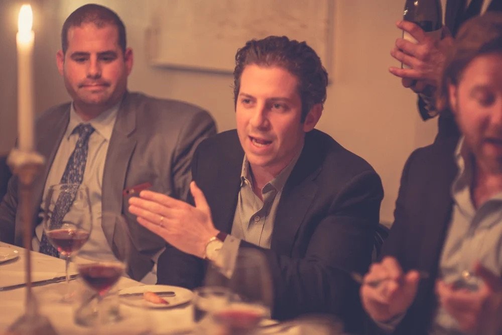 Man speaking at a dinner party with others seated around the table, some holding drinks, in a warm, dimly lit room.