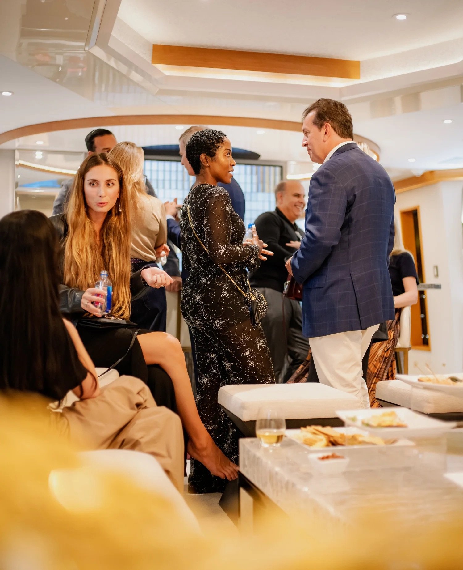 People socializing at a gathering in an indoor setting, with a woman in a black patterned dress and a man in a suit in the center, engaged in conversation. Others are seated or standing nearby, some holding drinks, with food and drinks on a table in the foreground.