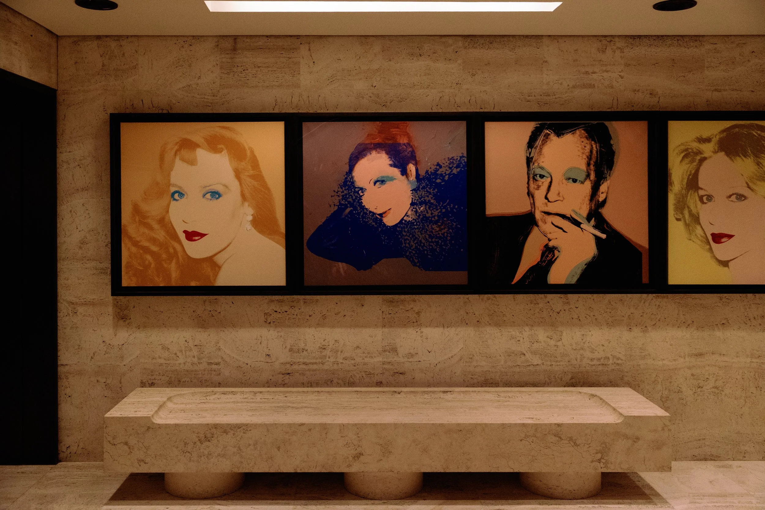 Four large framed pop art style portraits of women hanging on a beige stone wall, with a beige stone bench below.