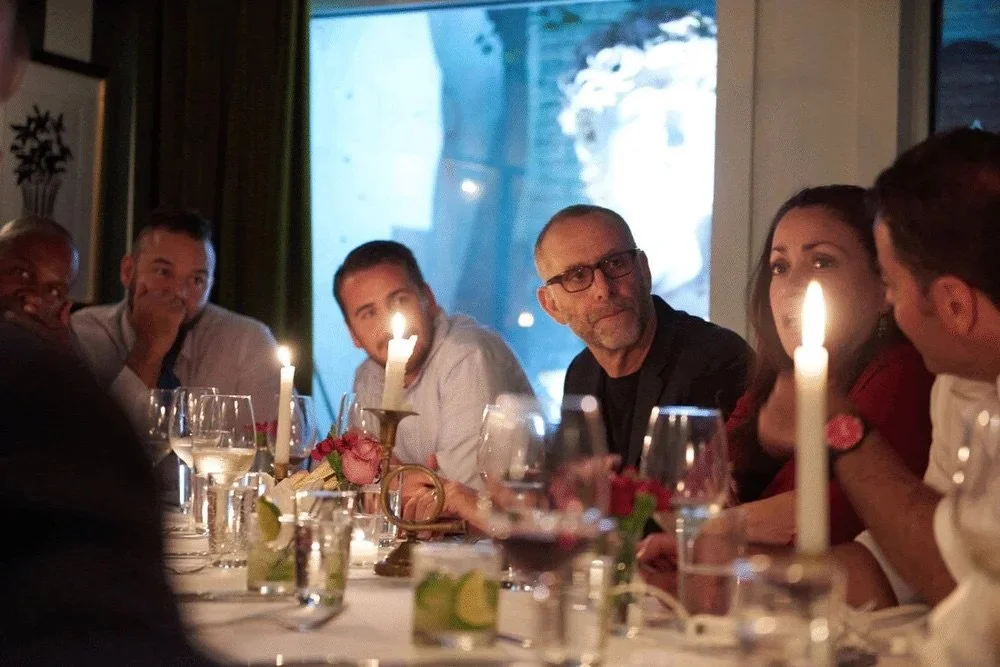 People sitting at a dinner table with candles and glasses, engaged in conversation in a dimly lit room.