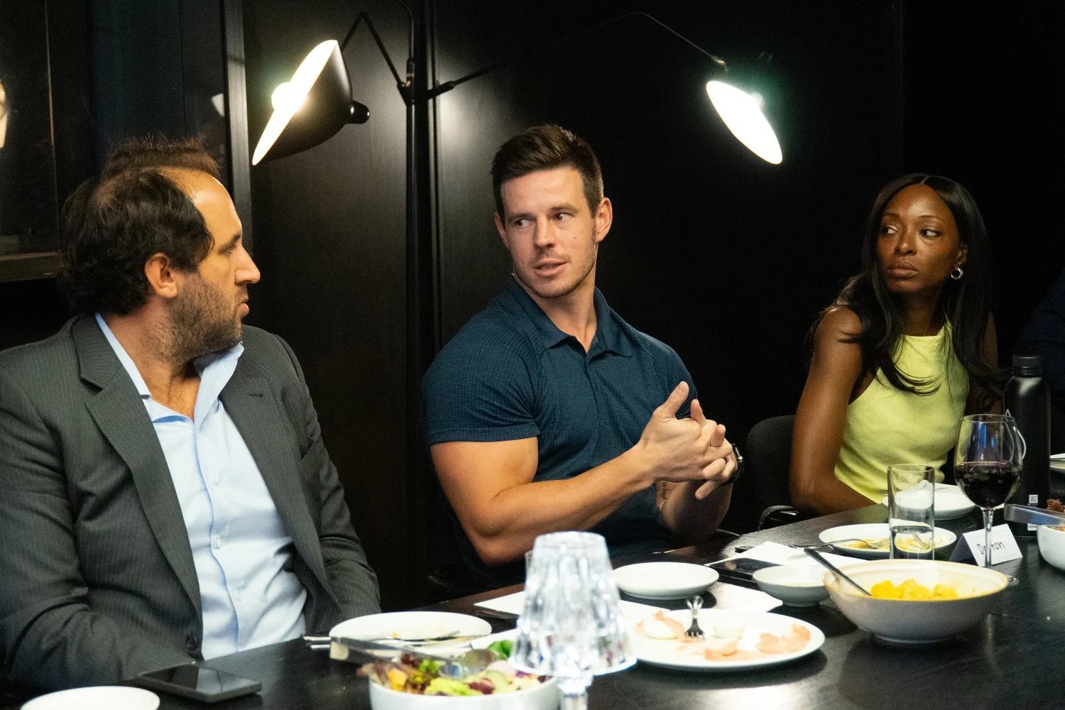 People sit at a dinner table in a dark room with black walls, with plates of food, wine, and water glasses, engaging in conversation.