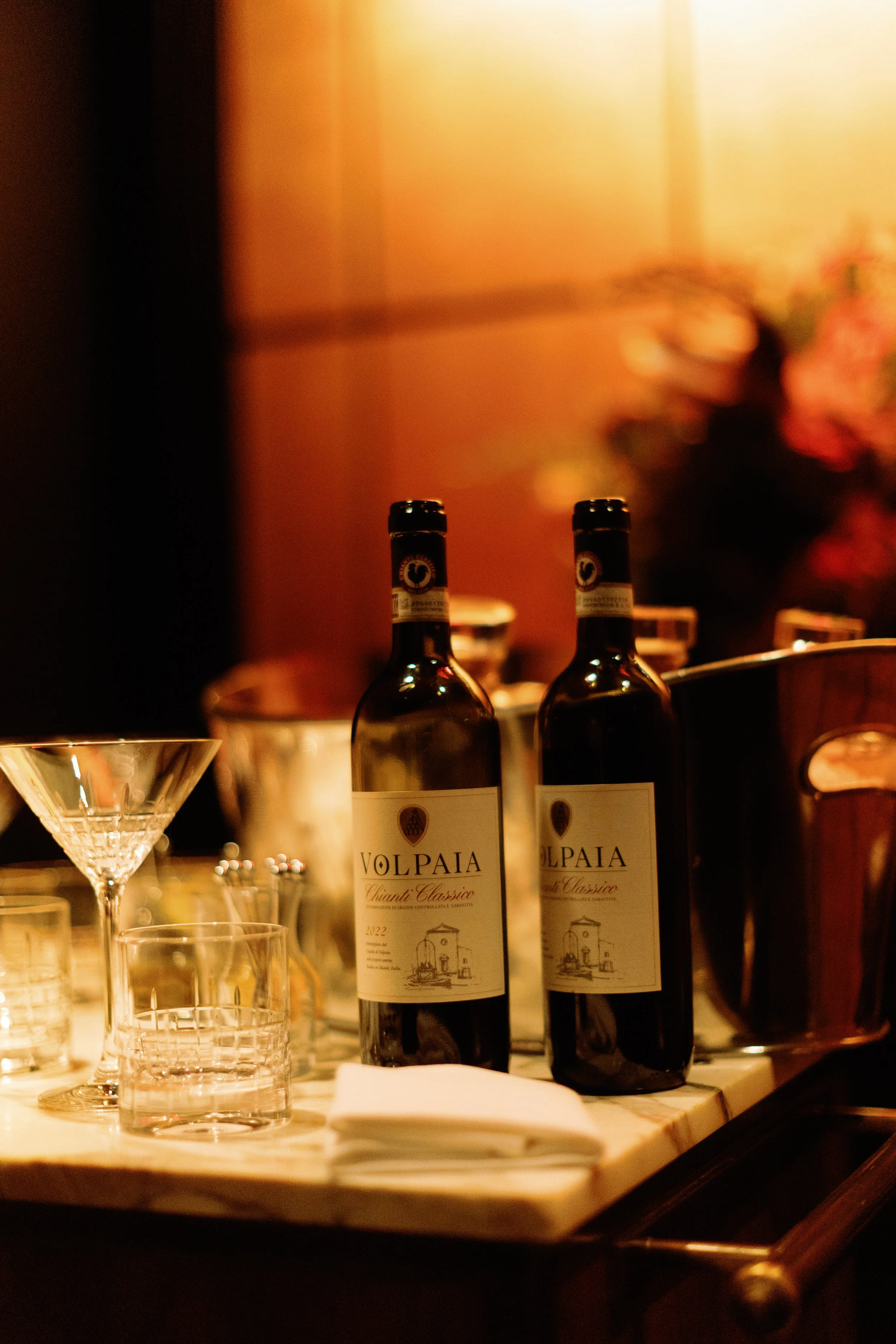 Two bottles of wine labeled 'Volpaia Chianti Classico' on a restaurant table, with glasses, a napkin, and blurred background of a warmly lit room with people.