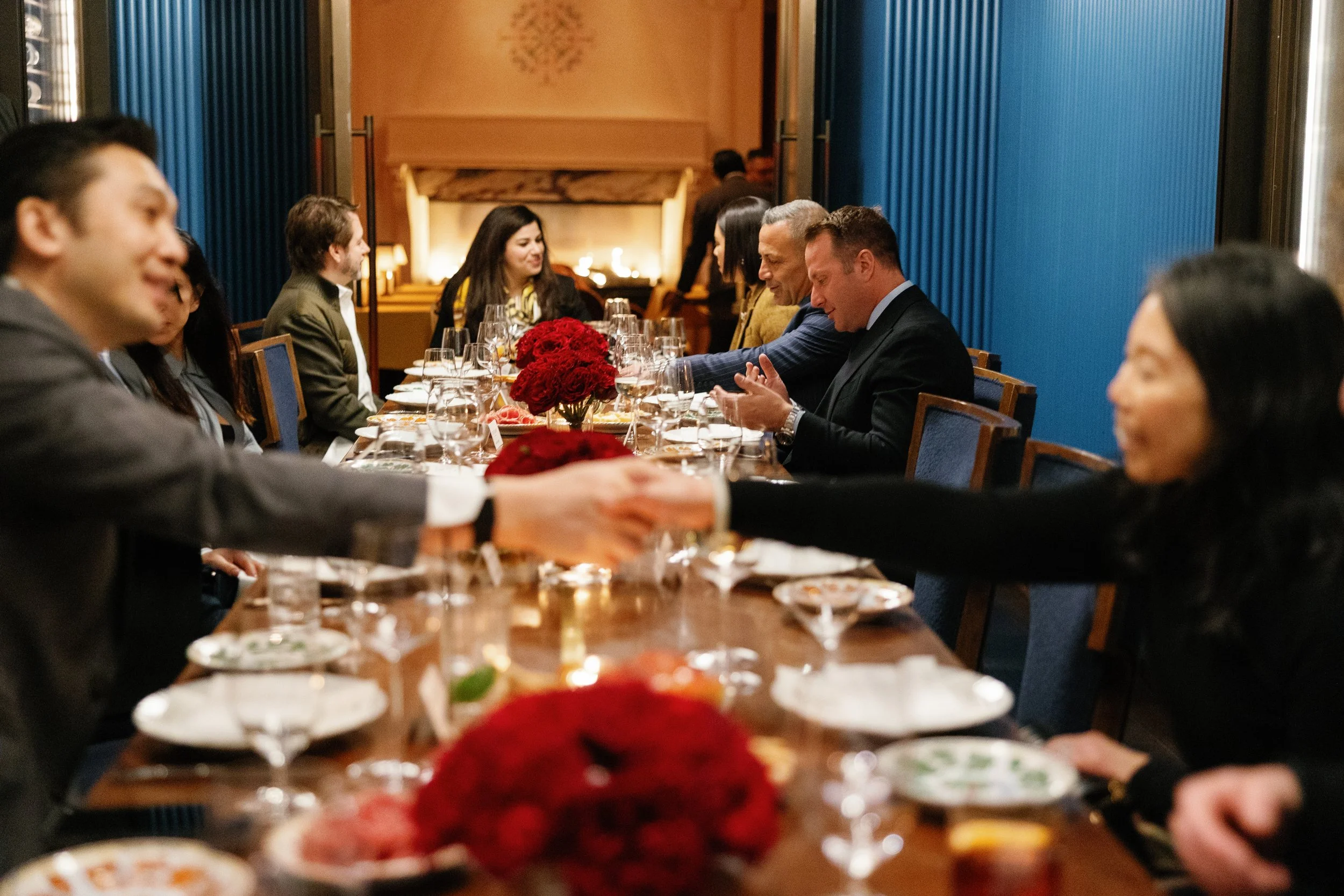 People seated at a long dining table with red floral centerpieces, having a meal in an elegant room with blue walls.