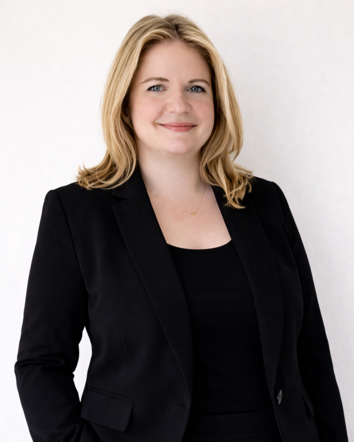 A professional woman with blonde, shoulder-length hair, wearing a black blazer and a delicate gold necklace, standing against a plain white background.