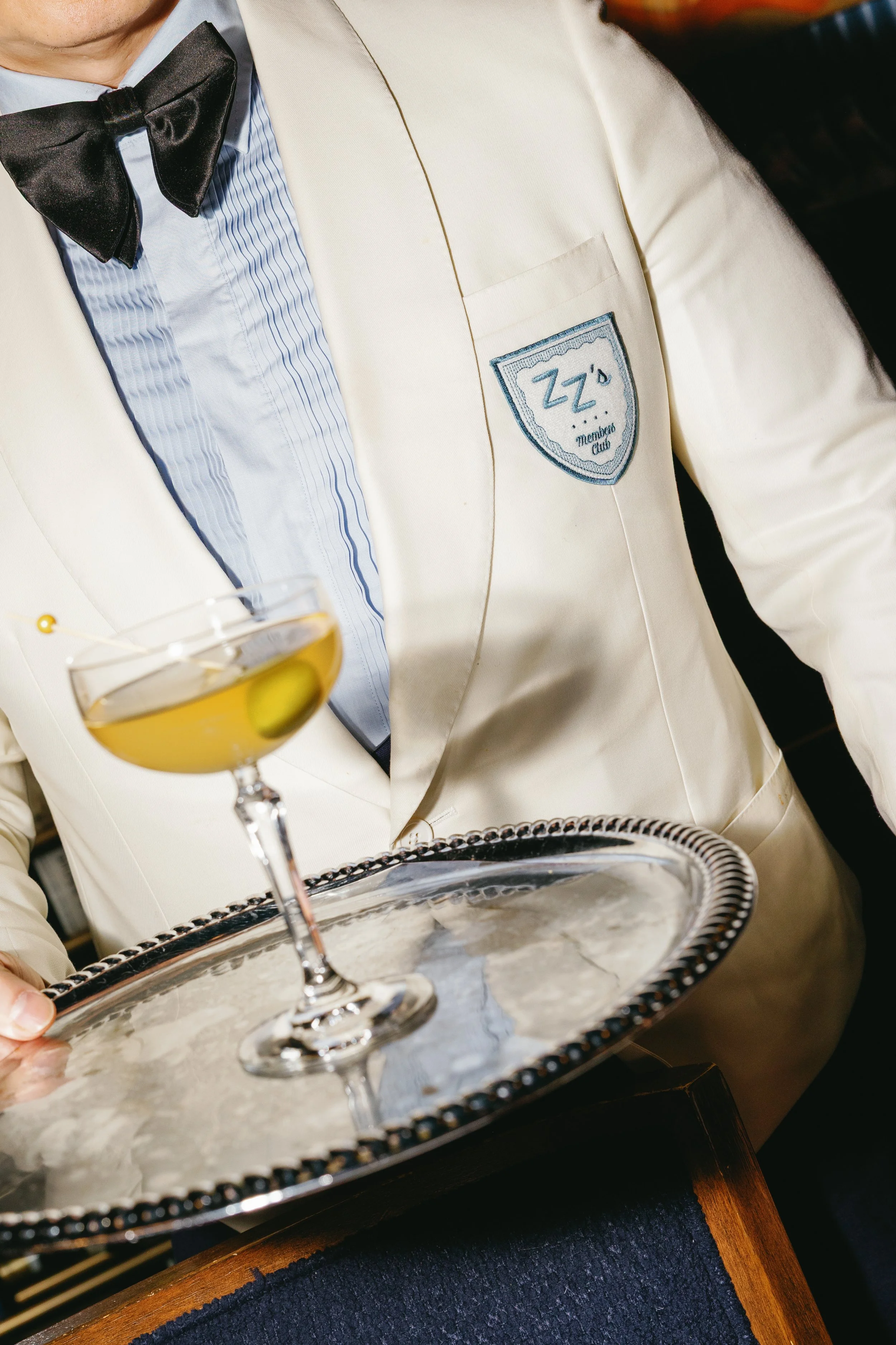Person dressed in a white tuxedo with a black bow tie and an embroidered badge that reads 'ZZ's Members Club,' holding a silver tray with drinks.