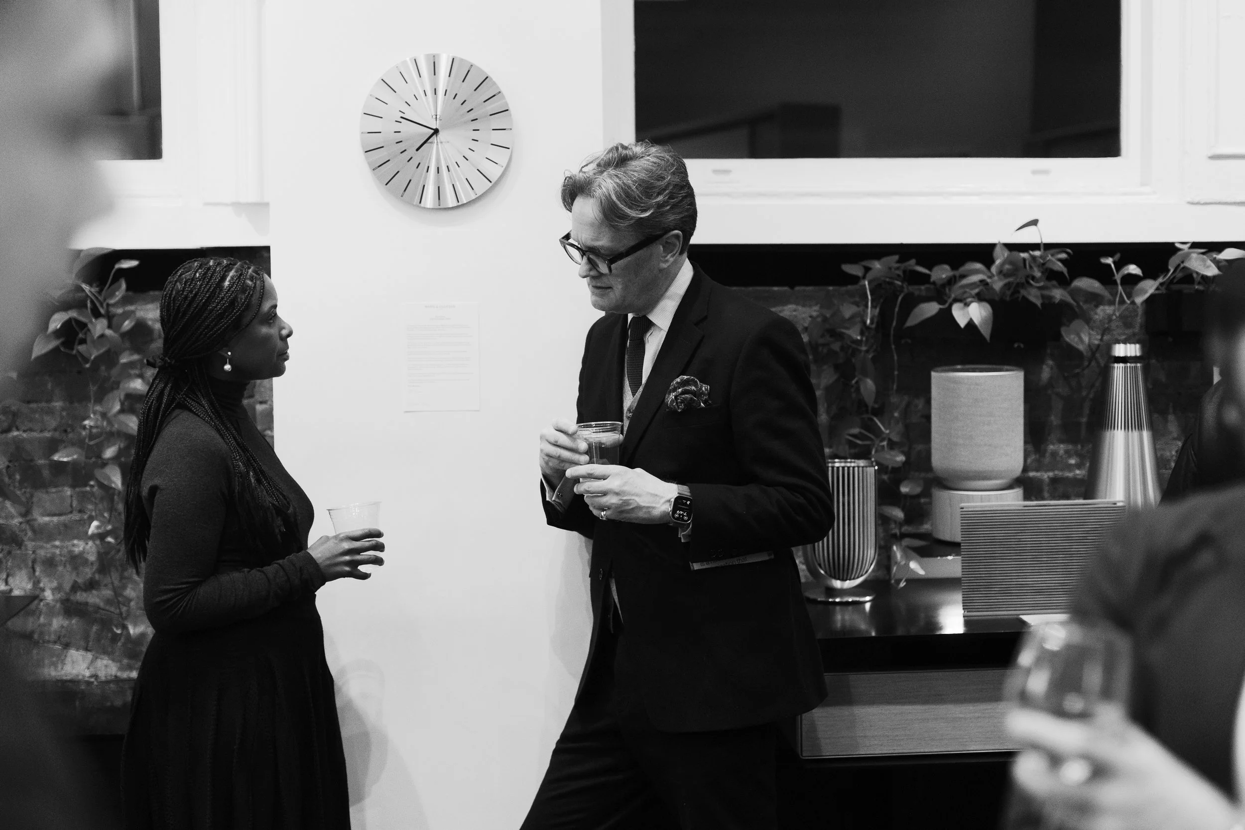 A woman and a man are having a conversation at an indoor gathering or event, with the woman holding a glass and the man holding a drink. The woman has braided hair and earrings, while the man is wearing glasses and a suit. There is a large clock on the wall and various decorative items, including plants and lamps, in the background.