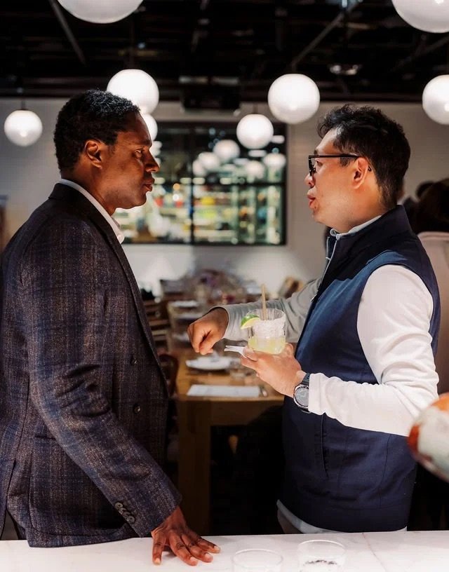 Two men talking in a restaurant; one holding a cocktail with lime, engaged in conversation with an expressive gesture.