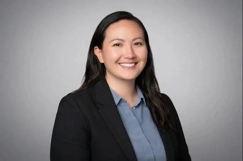 A woman with long dark hair smiling, wearing a black blazer and a blue collared shirt, against a plain gray background.