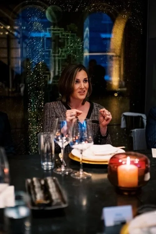 A woman with shoulder-length brown hair sitting at a dining table with wine glasses, a lit candle in a glass holder, and plates, with a rainy cityscape visible through the window behind her.