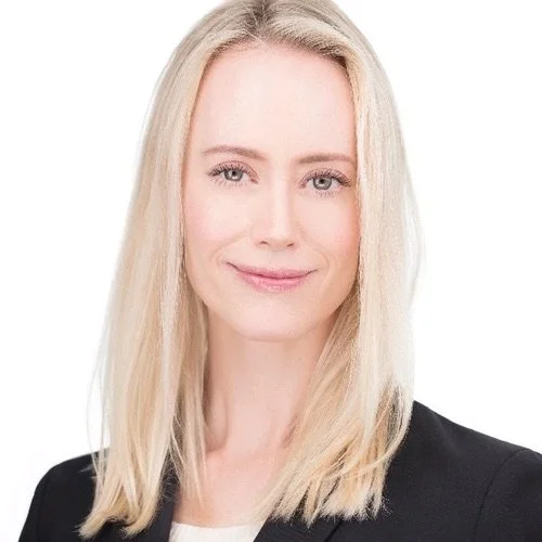 A portrait of a woman with long blonde hair, wearing a black blazer, smiling softly against a plain white background.
