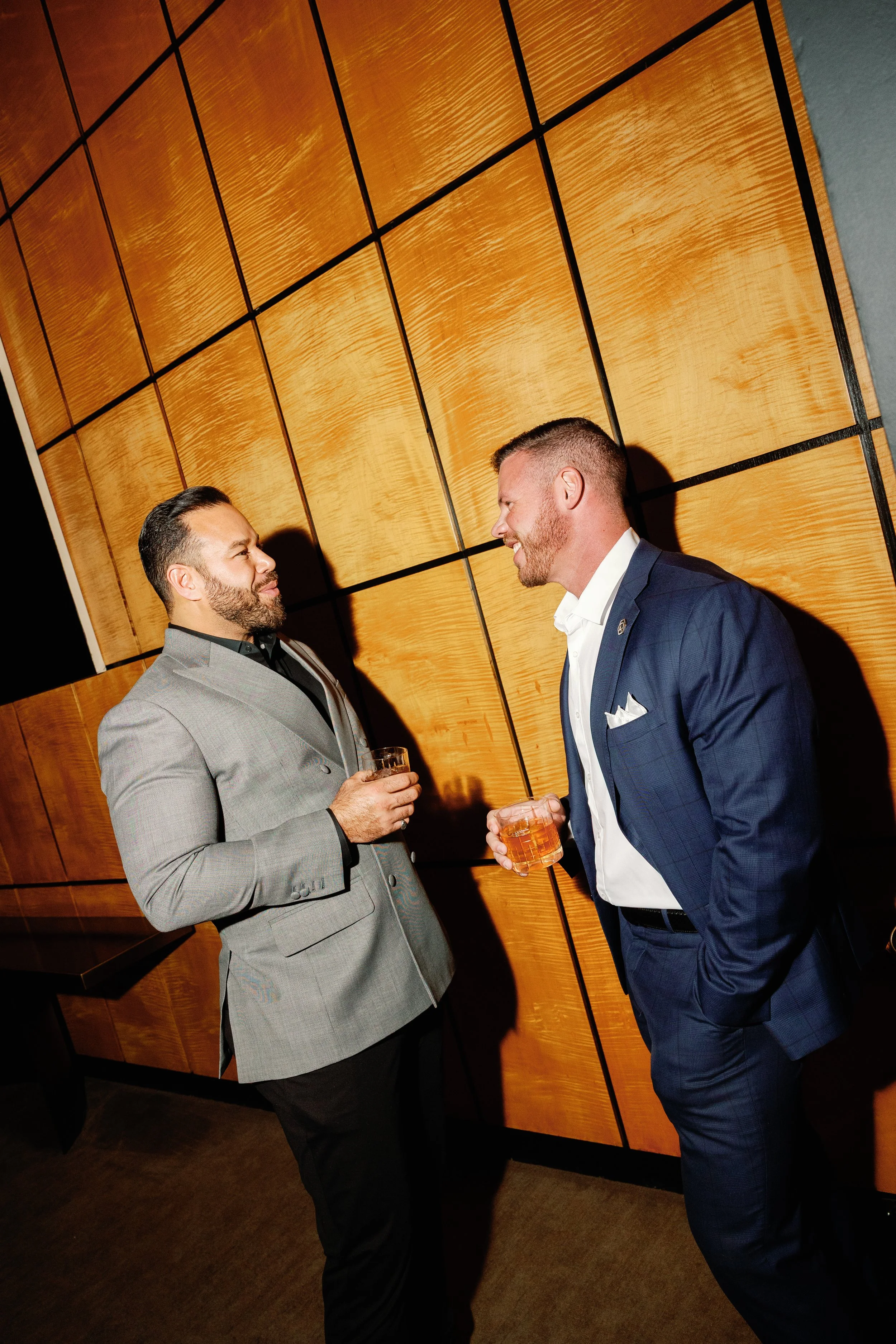Two men in suits having a conversation and smiling at each other at a social event, holding glasses of drinks, standing against a wooden-paneled wall.