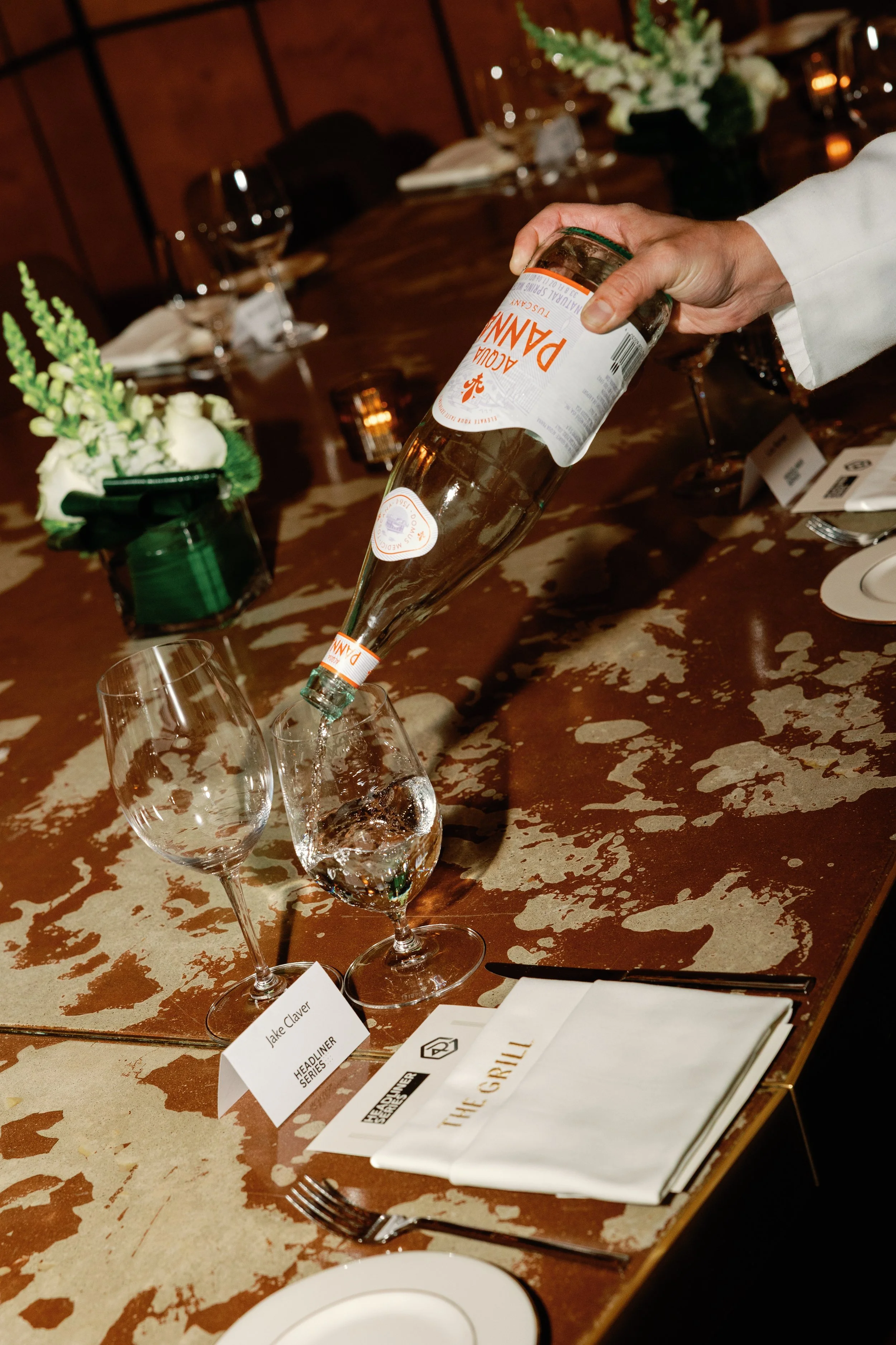 Person pouring champagne into wine glasses on a wooden dining table with floral centerpieces and place cards.
